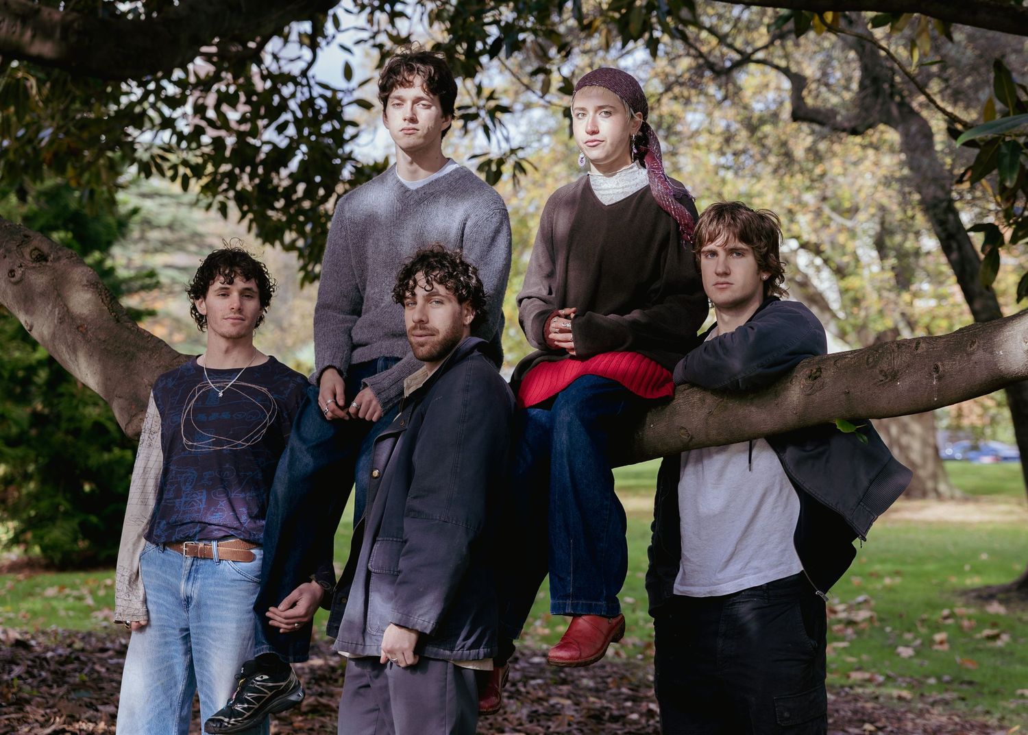 Five young people posing outdoors among trees, with some sitting on a branch in a lush green park setting.