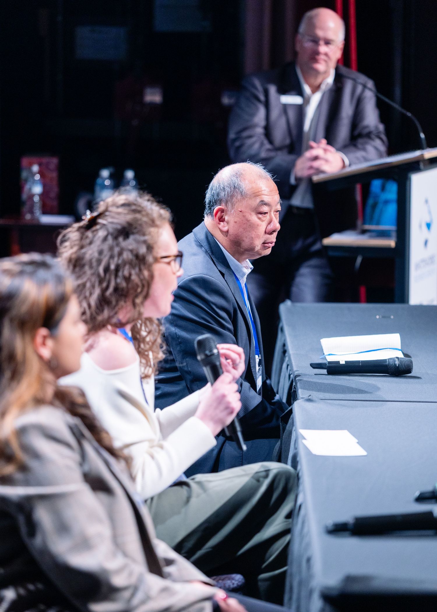 Panel of speakers seated at table during conference with moderator standing at podium in background.