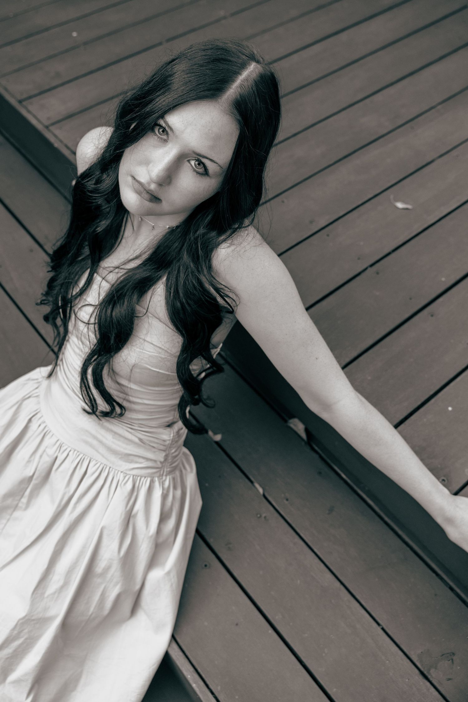 Black and white photo of a young woman with long dark hair in a white dress sitting on wooden deck steps.