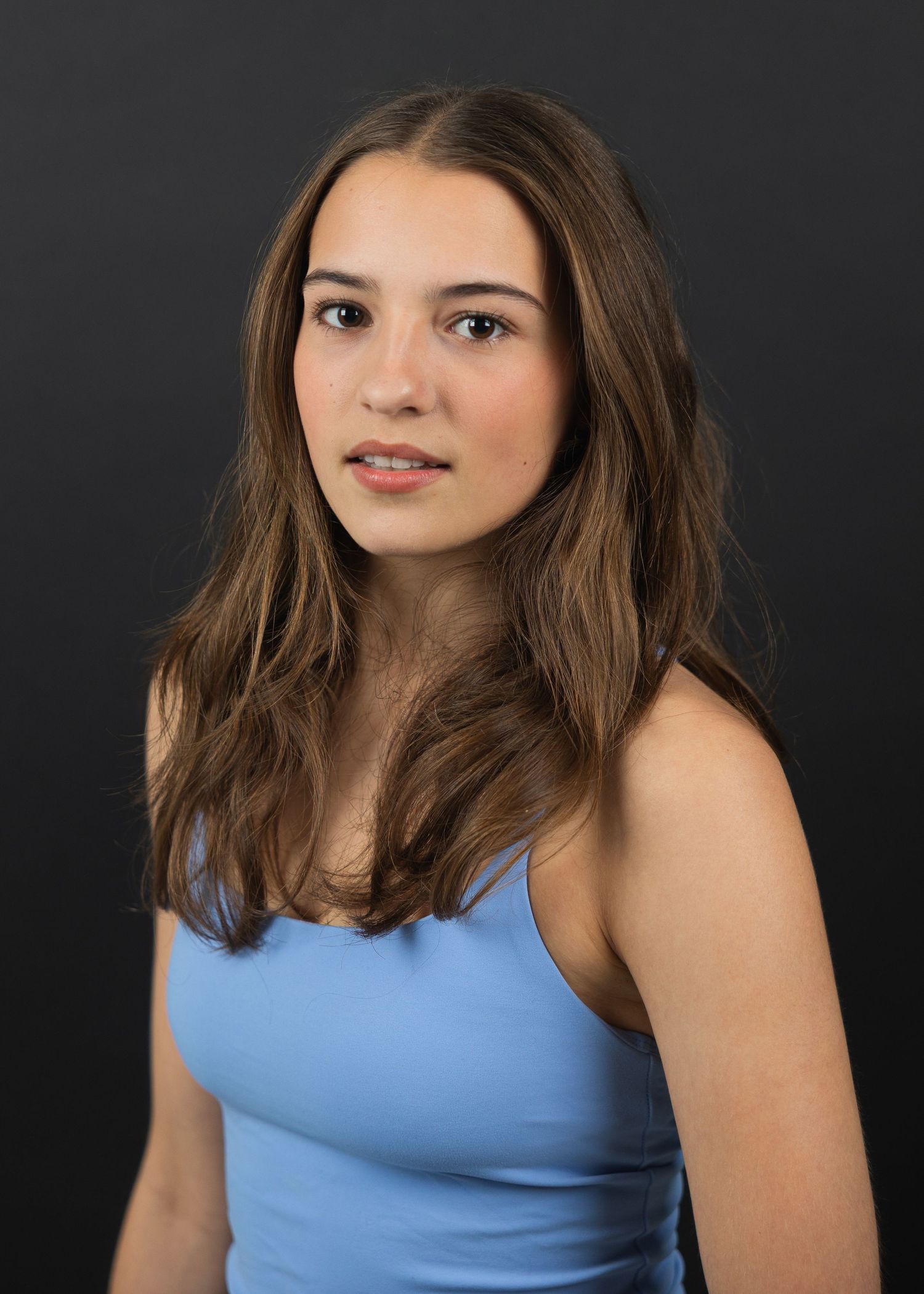 Young woman with long brown hair wearing a light blue tank top poses against a dark gray background.