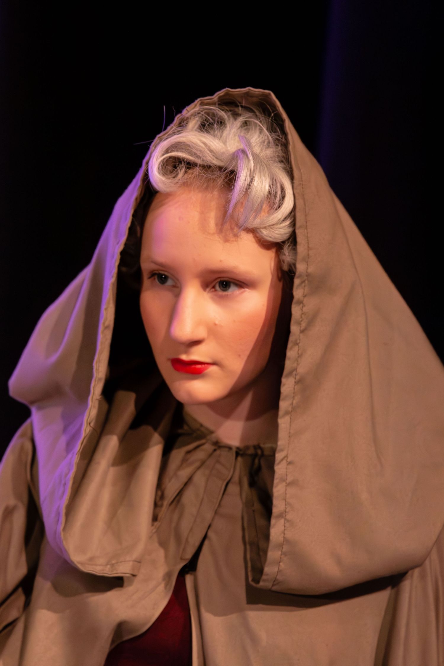 Young woman in a tan hooded cloak with silver curled hair and red lips poses dramatically on a dark stage.