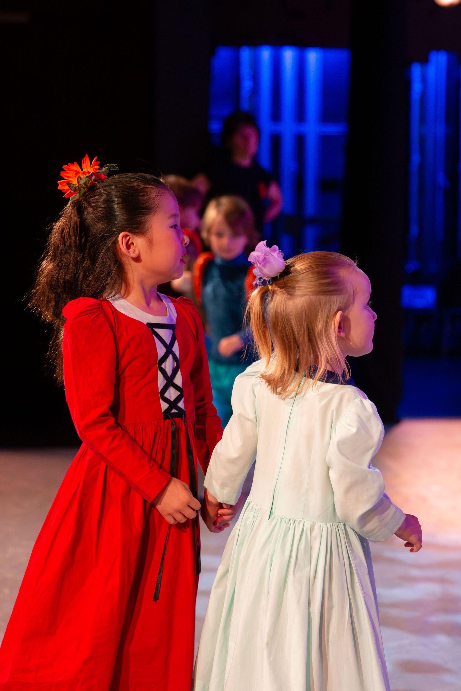 Two young girls in red and white dresses hold hands on stage during a children's theater performance.