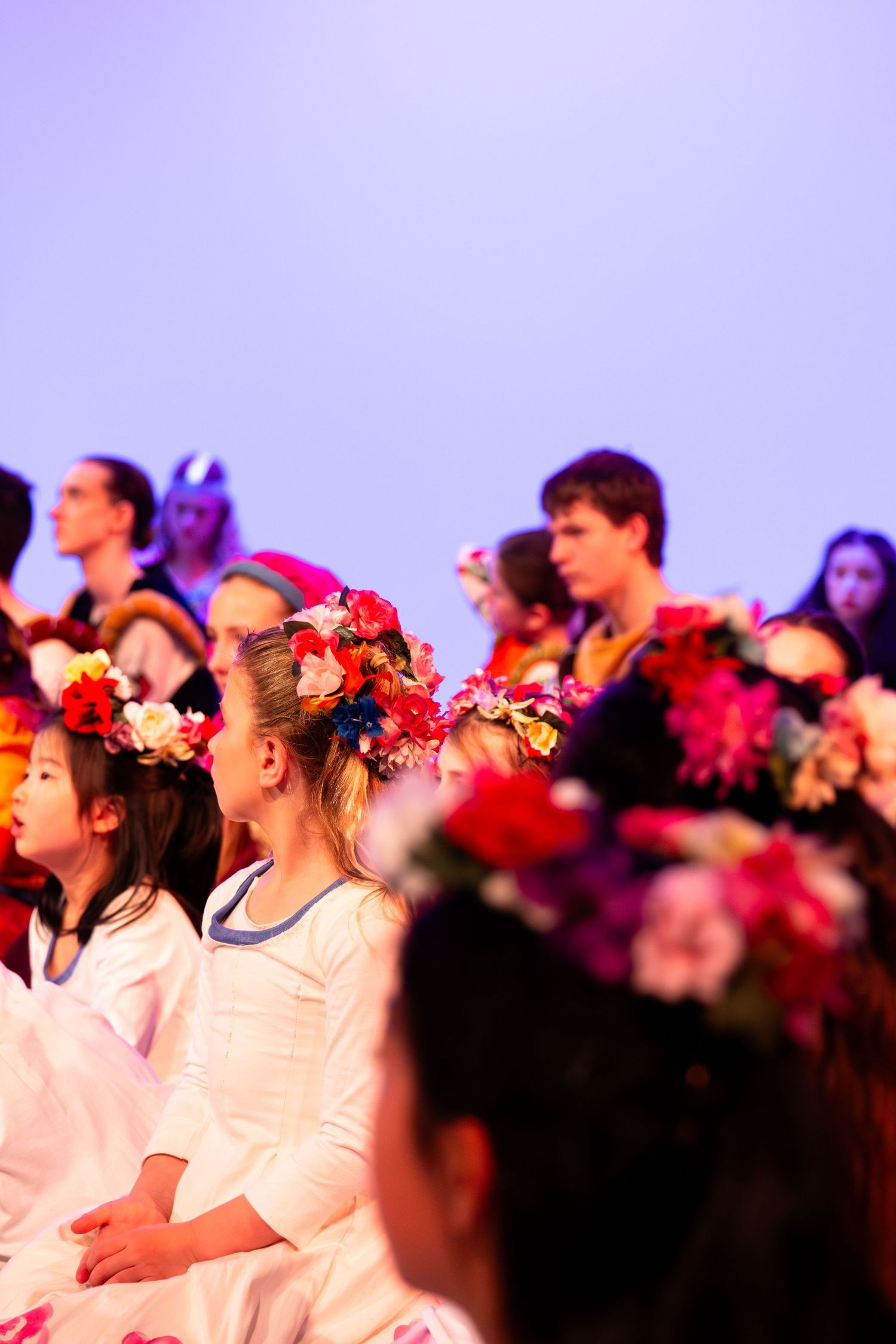Children and performers wearing colorful flower crowns sit together on stage during a festive performance or recital.