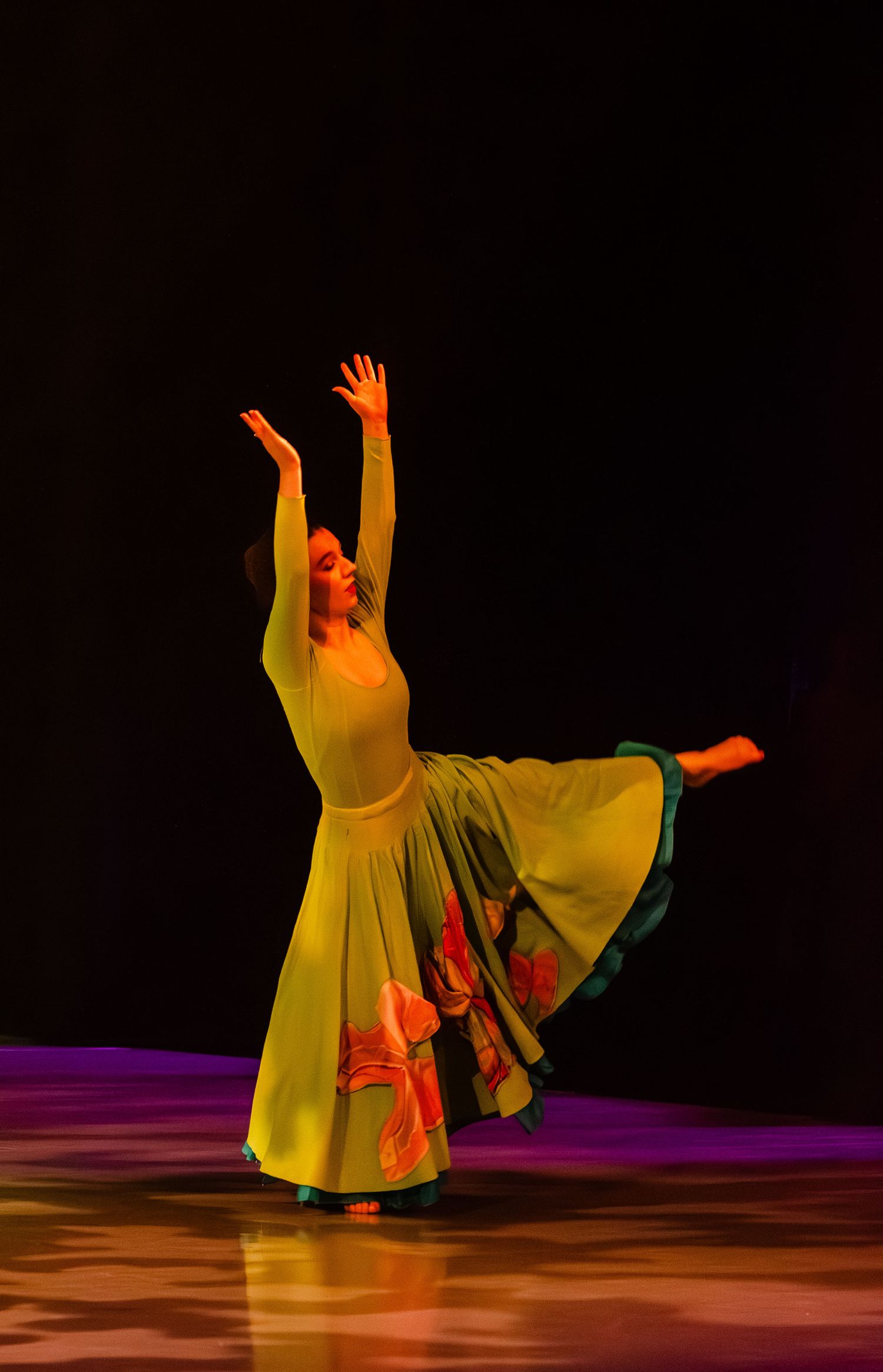 A dancer in flowing yellow robes performs an expressive modern dance sequence on a darkened stage with colored lighting.