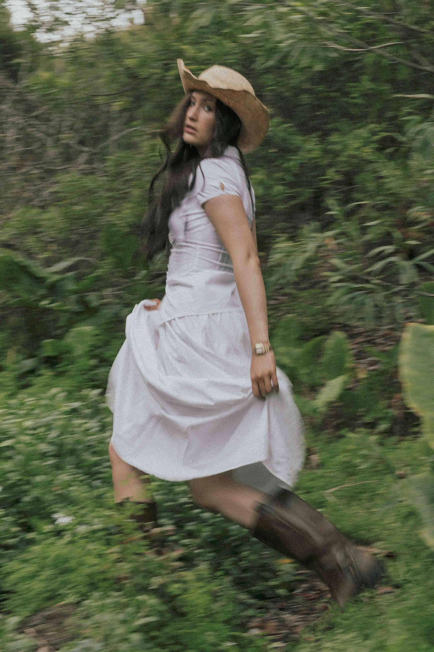 Woman in white dress, cowboy hat, and boots running through lush green forest, motion blur effect.