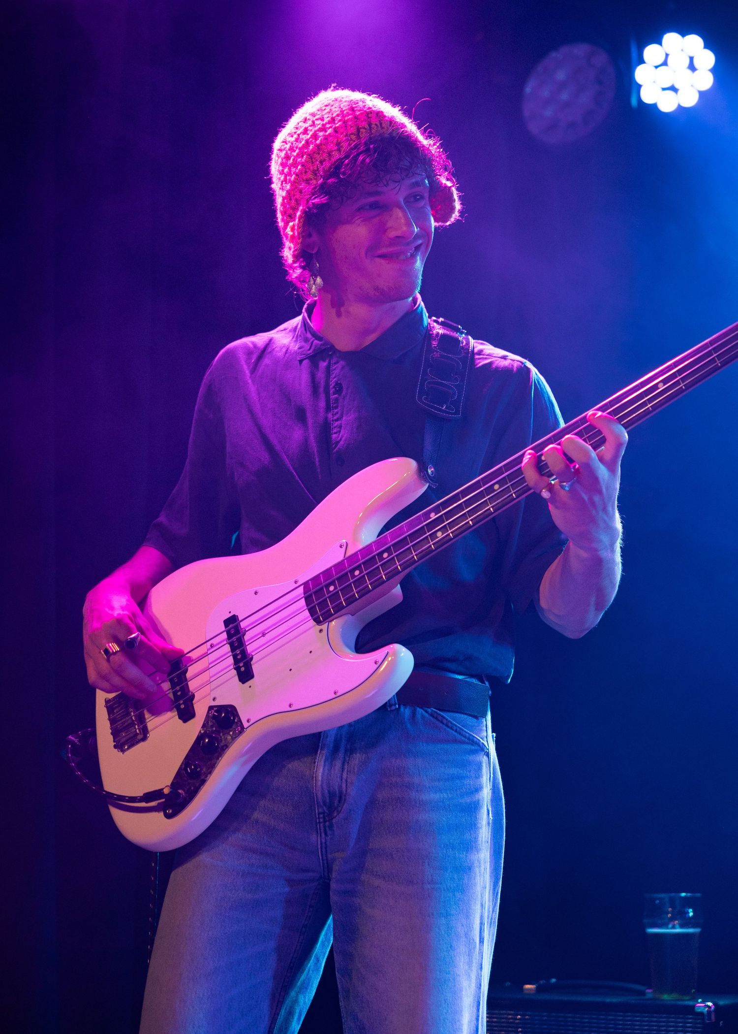 Young musician playing white bass guitar on stage under purple lights wearing a pink knit beanie.