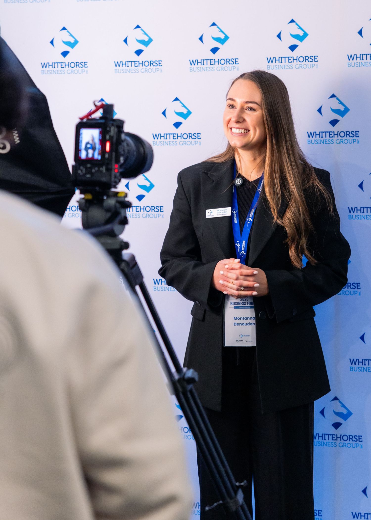 A smiling woman in a black blazer with a name badge poses in front of a Whitehorse Business Group branded backdrop.
