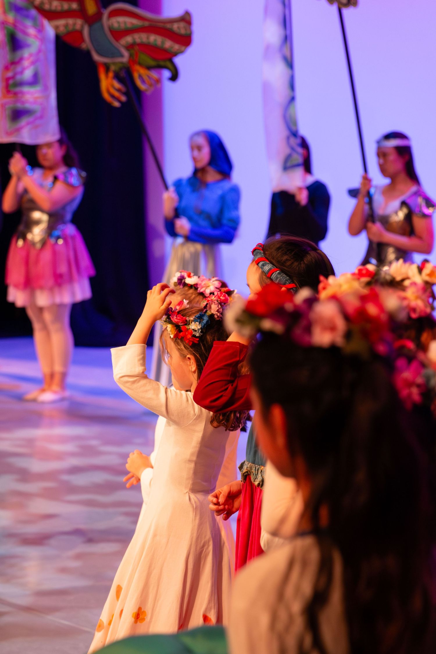 Girls in floral crowns perform a cultural dance on stage during a colorful theatrical performance.