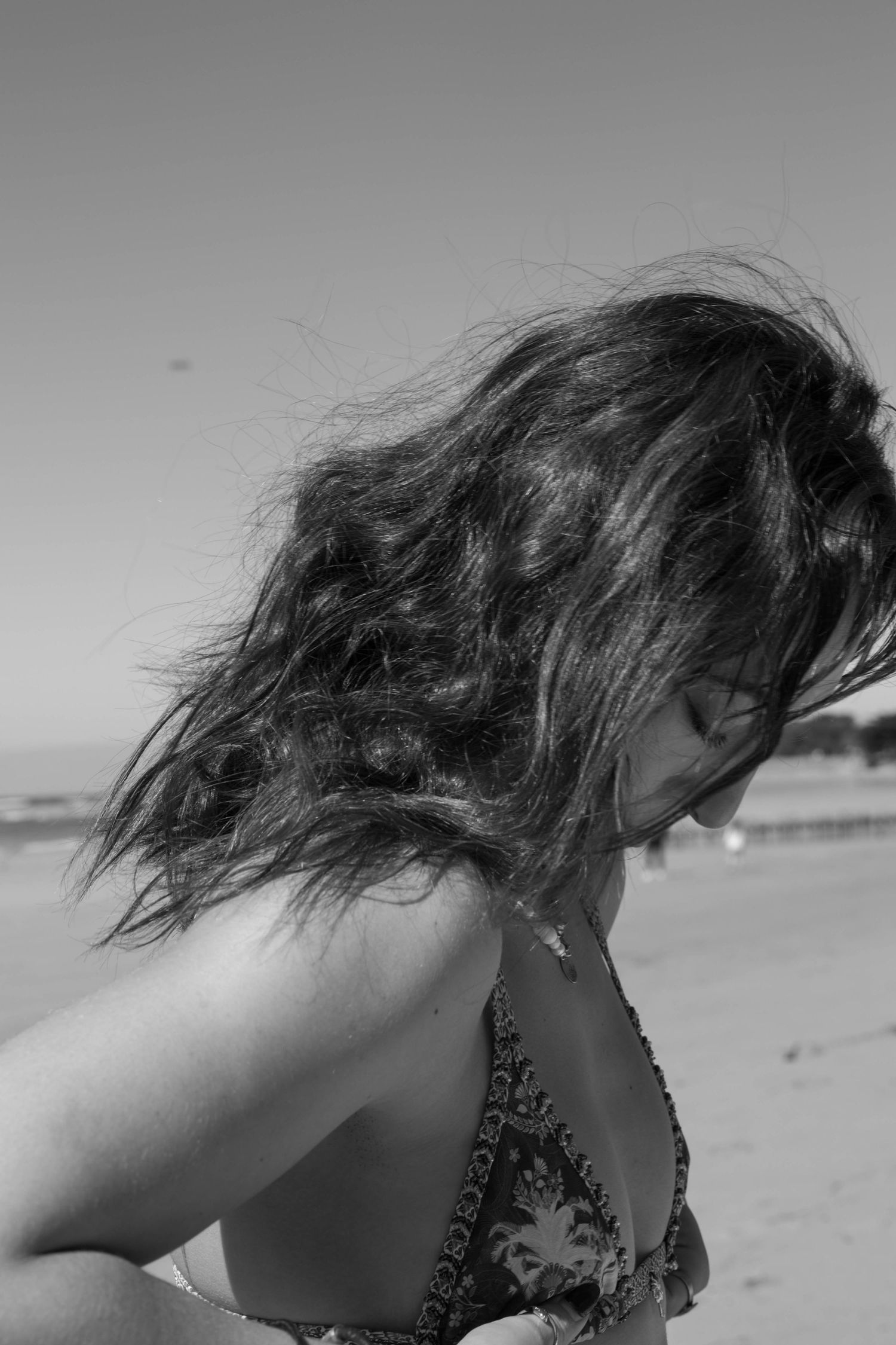 Black and white photo of a woman with windswept hair on a beach wearing a patterned bikini top.