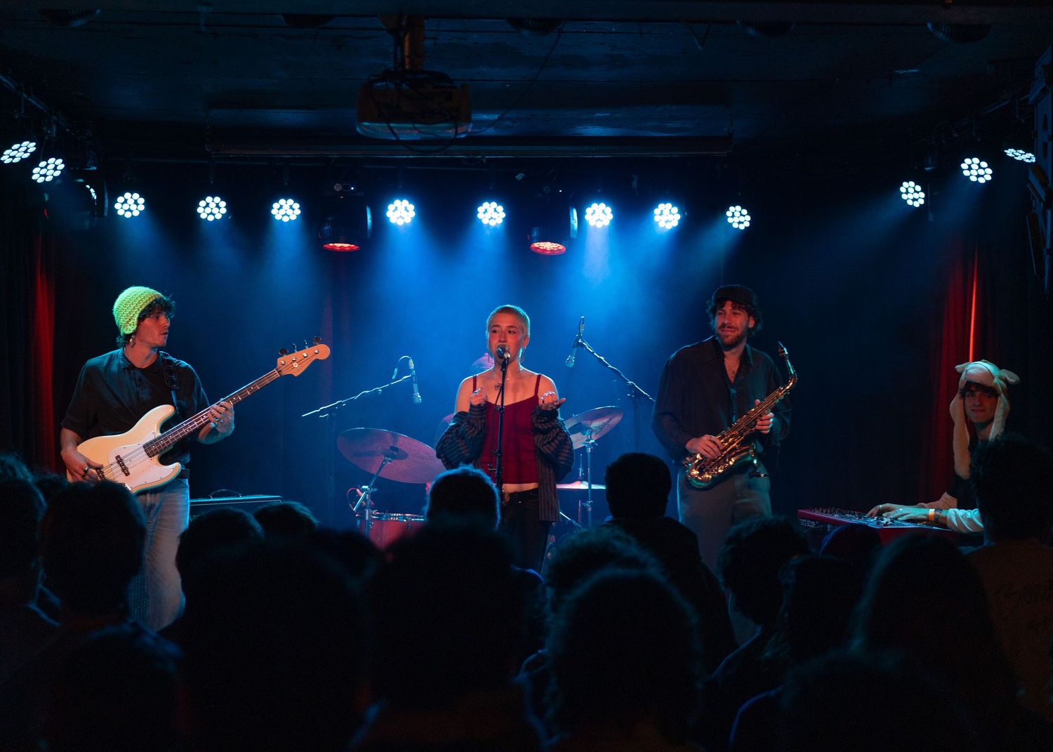 Live band performing on stage with colorful blue and red lights, guitarist, vocalist, saxophonist, and drummer.