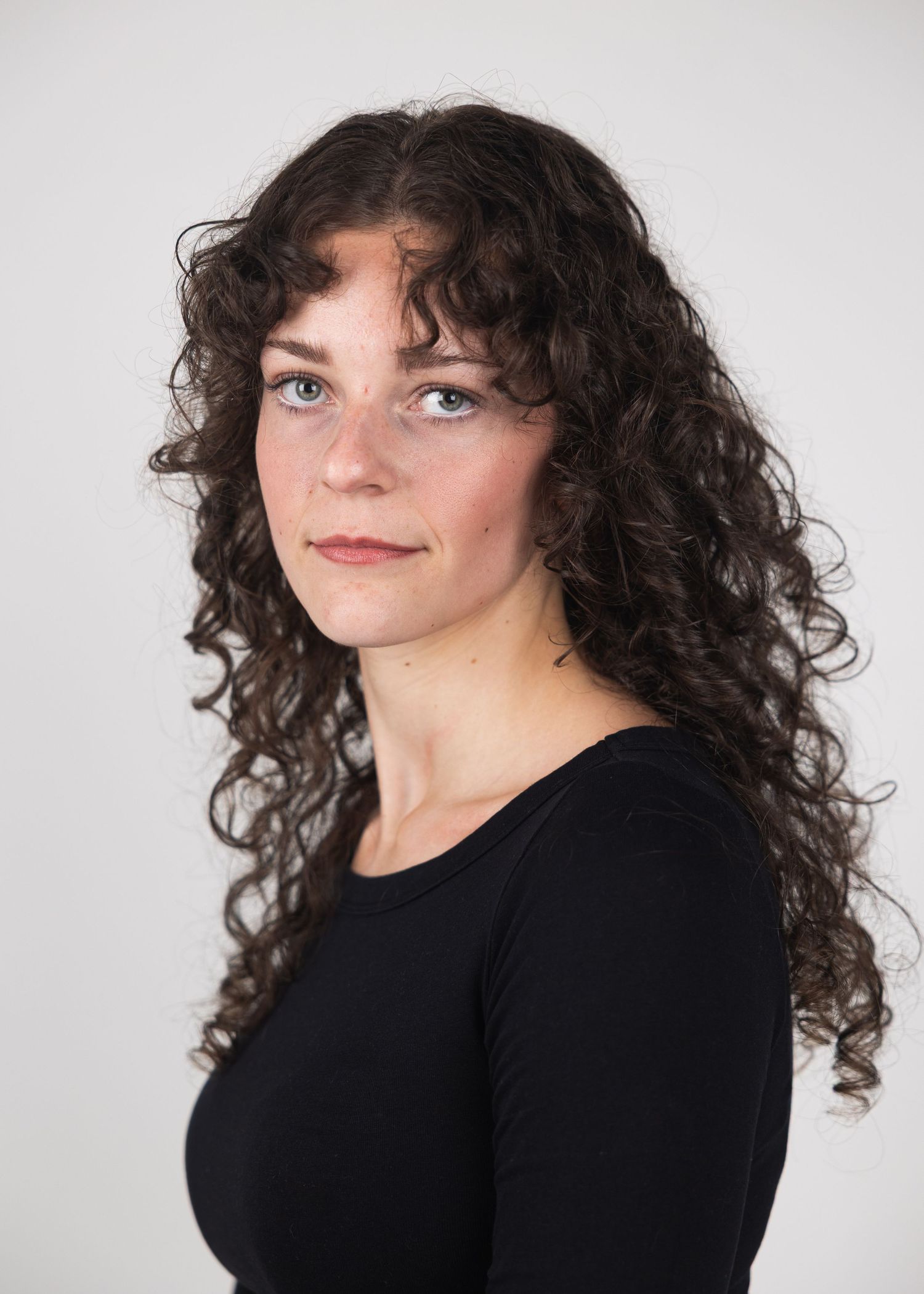 Young woman with long curly dark hair and blue eyes wearing a black top against a light gray background.