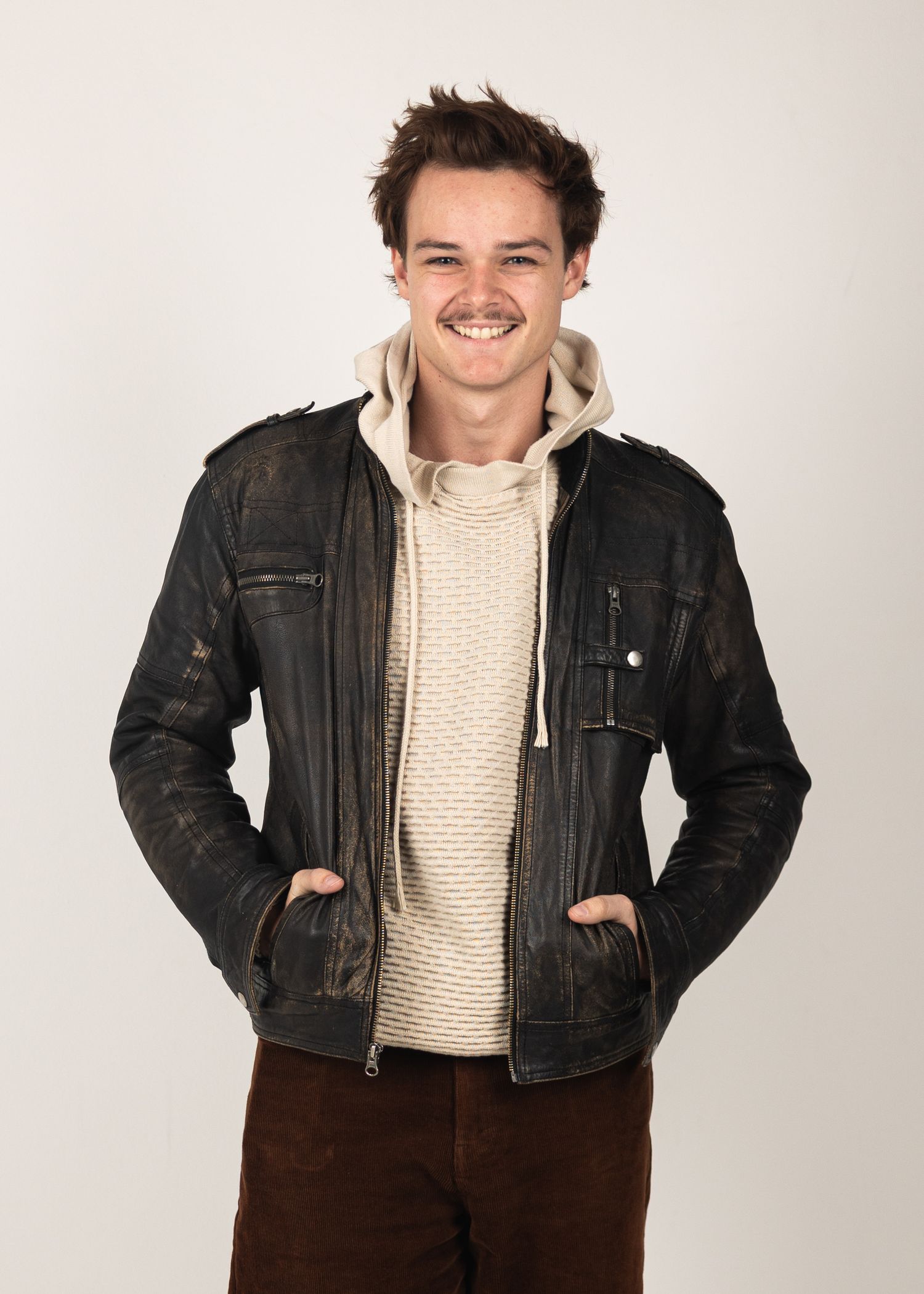 Young man smiling in a distressed black leather jacket over a cream hoodie and brown pants.