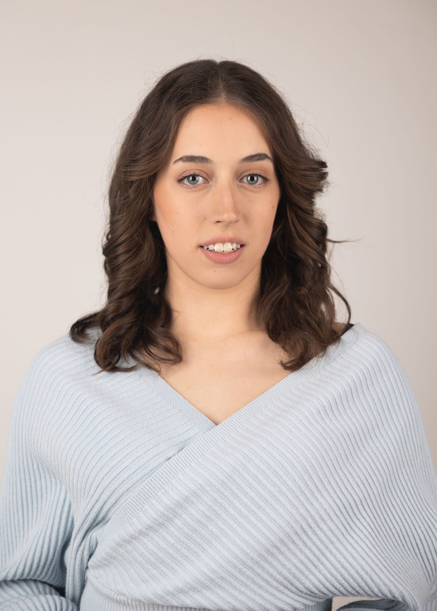 Young woman with blue eyes and wavy brown hair wearing a light blue wrap top, posing for a professional headshot.