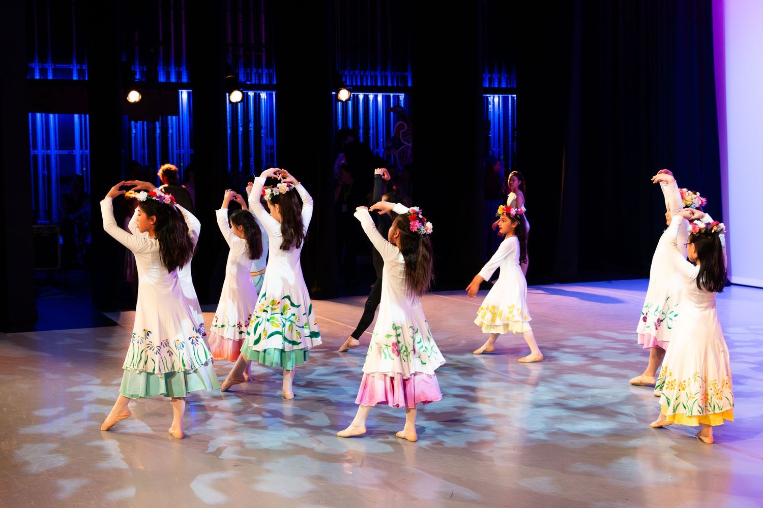 Young girls in white floral dresses perform a graceful folk dance on a lit stage with colorful lighting.