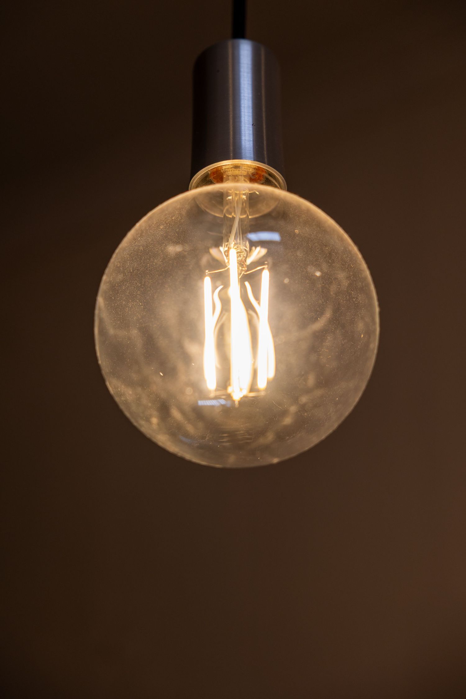 Vintage Edison globe light bulb with glowing filament hanging from ceiling against dark brown background.