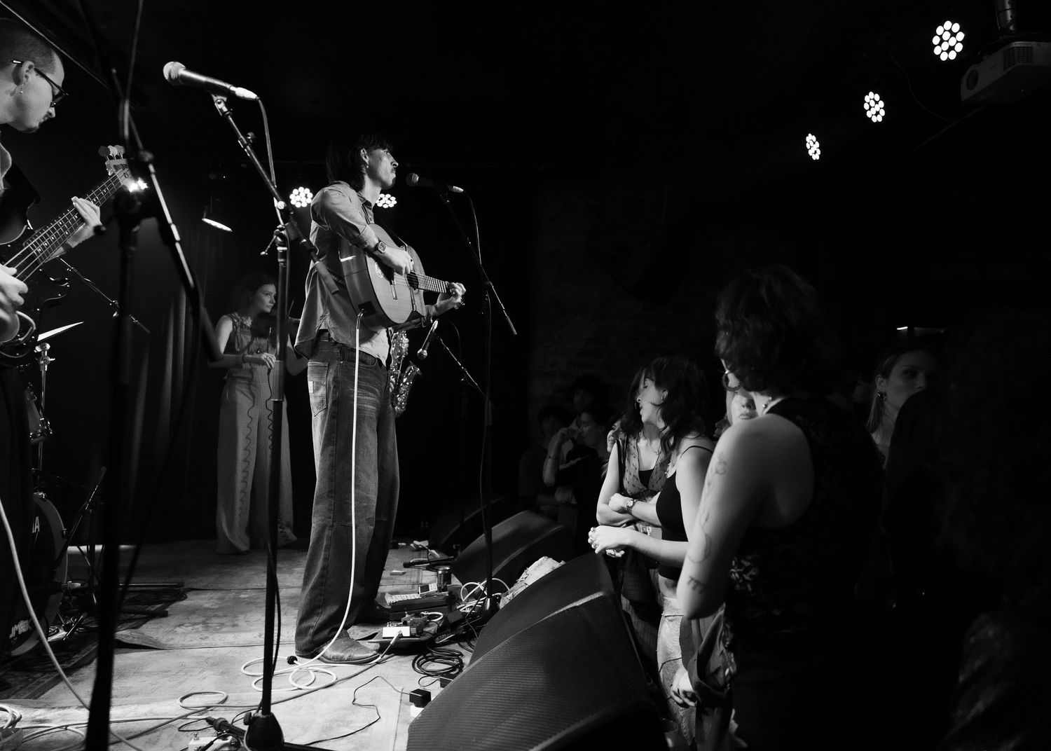 Black and white photo of a live band performance on stage with guitarist, microphones, and audience watching closely.