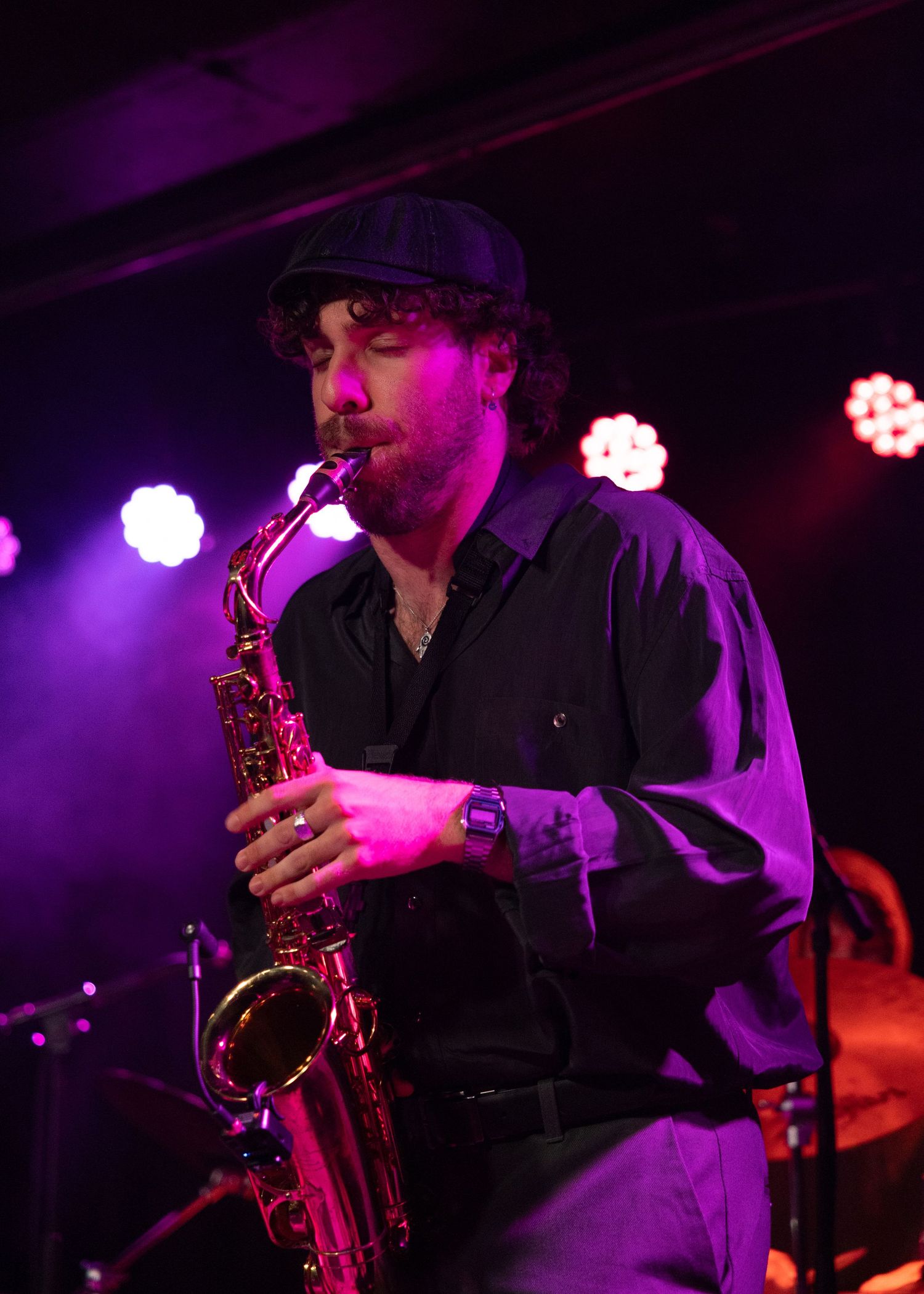 A male musician plays saxophone on a dimly lit stage with purple lighting during a live jazz performance.