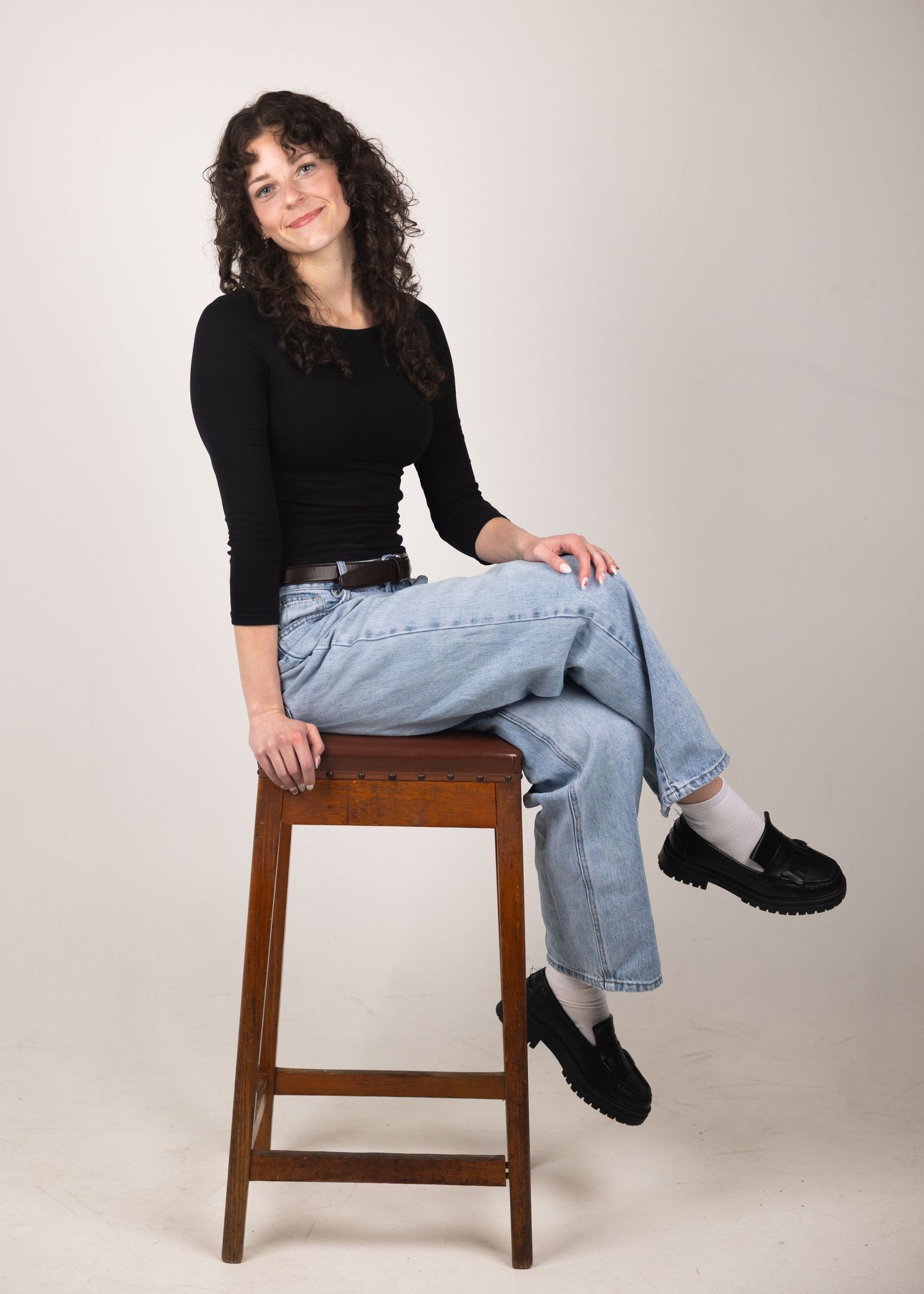 Young woman with curly hair sits on wooden stool wearing black top, light jeans, and black loafers against neutral background.