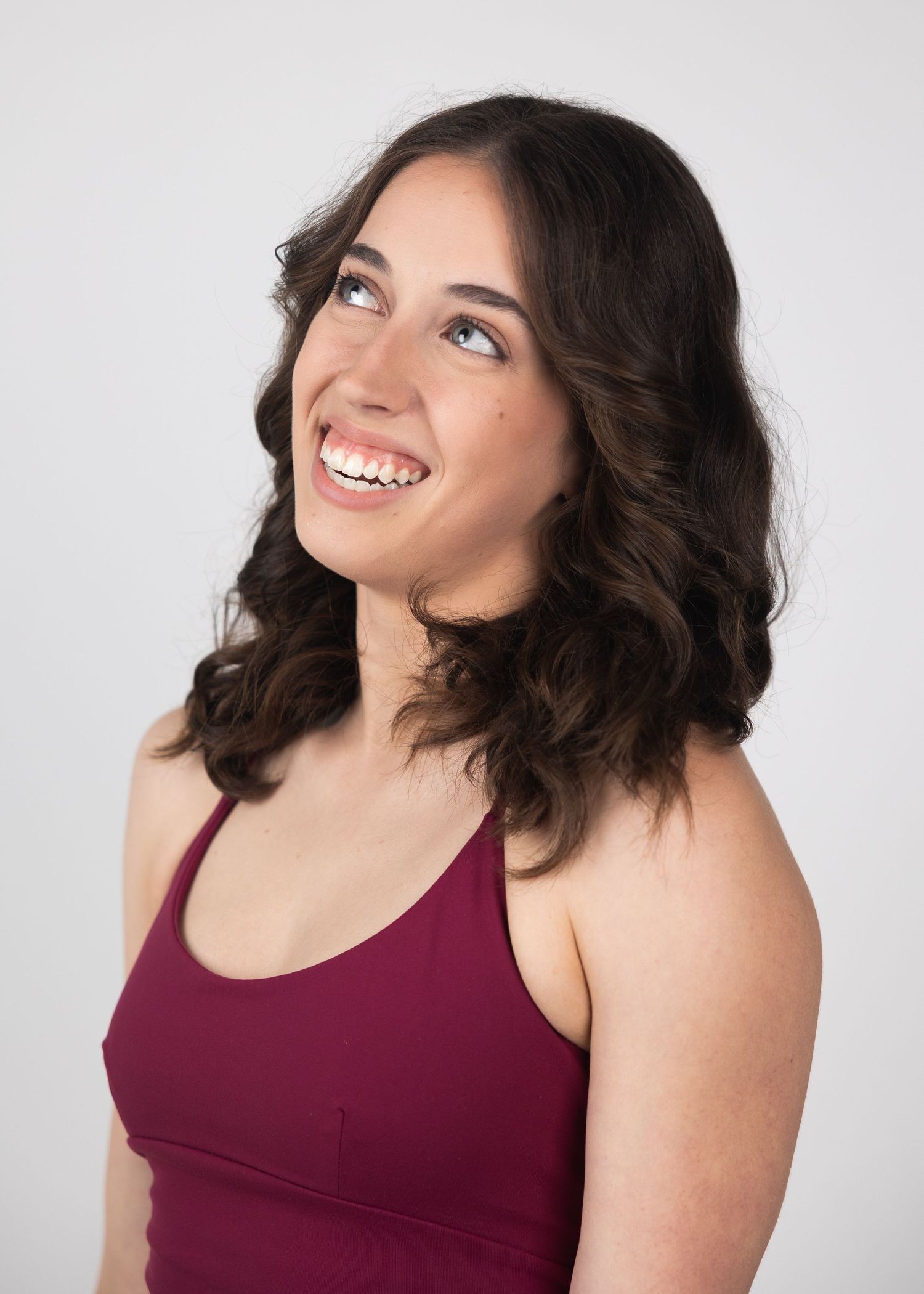 Smiling young woman with dark wavy hair wearing maroon tank top looking up against white background.