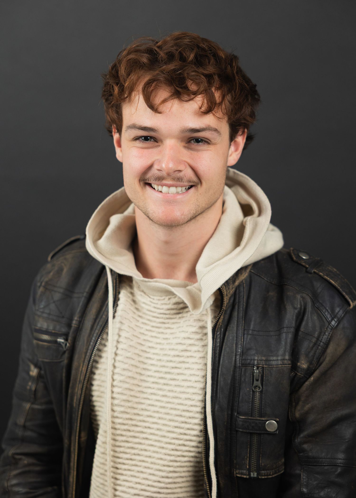 Smiling young man with curly hair wearing a leather jacket and cream hoodie sweater.