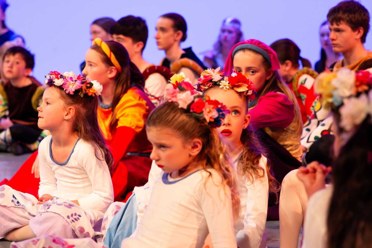 Children in floral headbands sit together during a colorful stage performance or school recital.