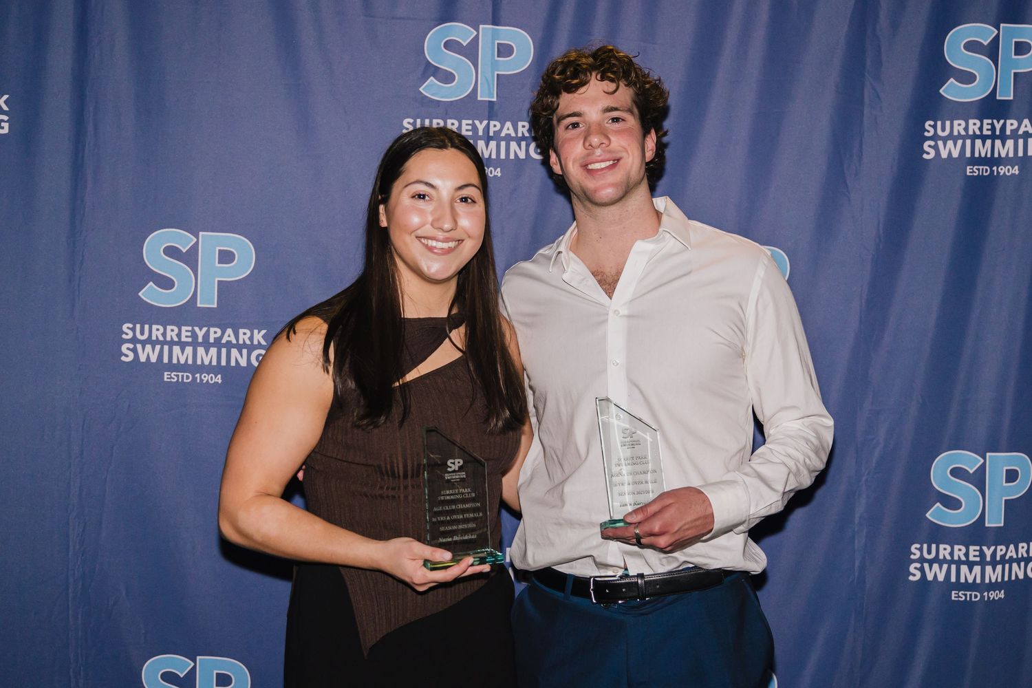 Two swimmers proudly hold crystal awards at Surrey Park Swimming Club annual ceremony against branded blue backdrop.