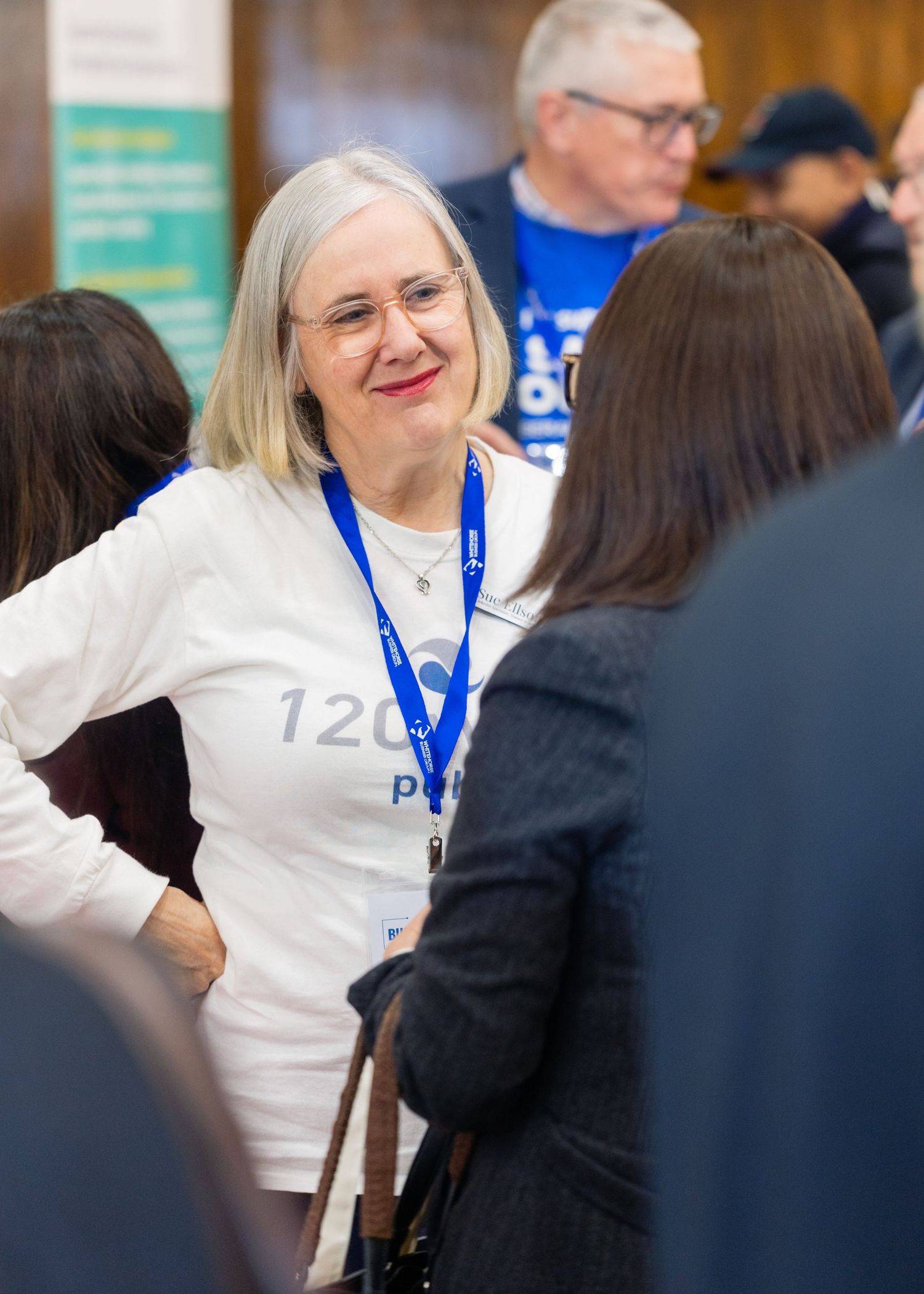 Woman with glasses and blue lanyard smiling at a networking event, wearing a white shirt with text.