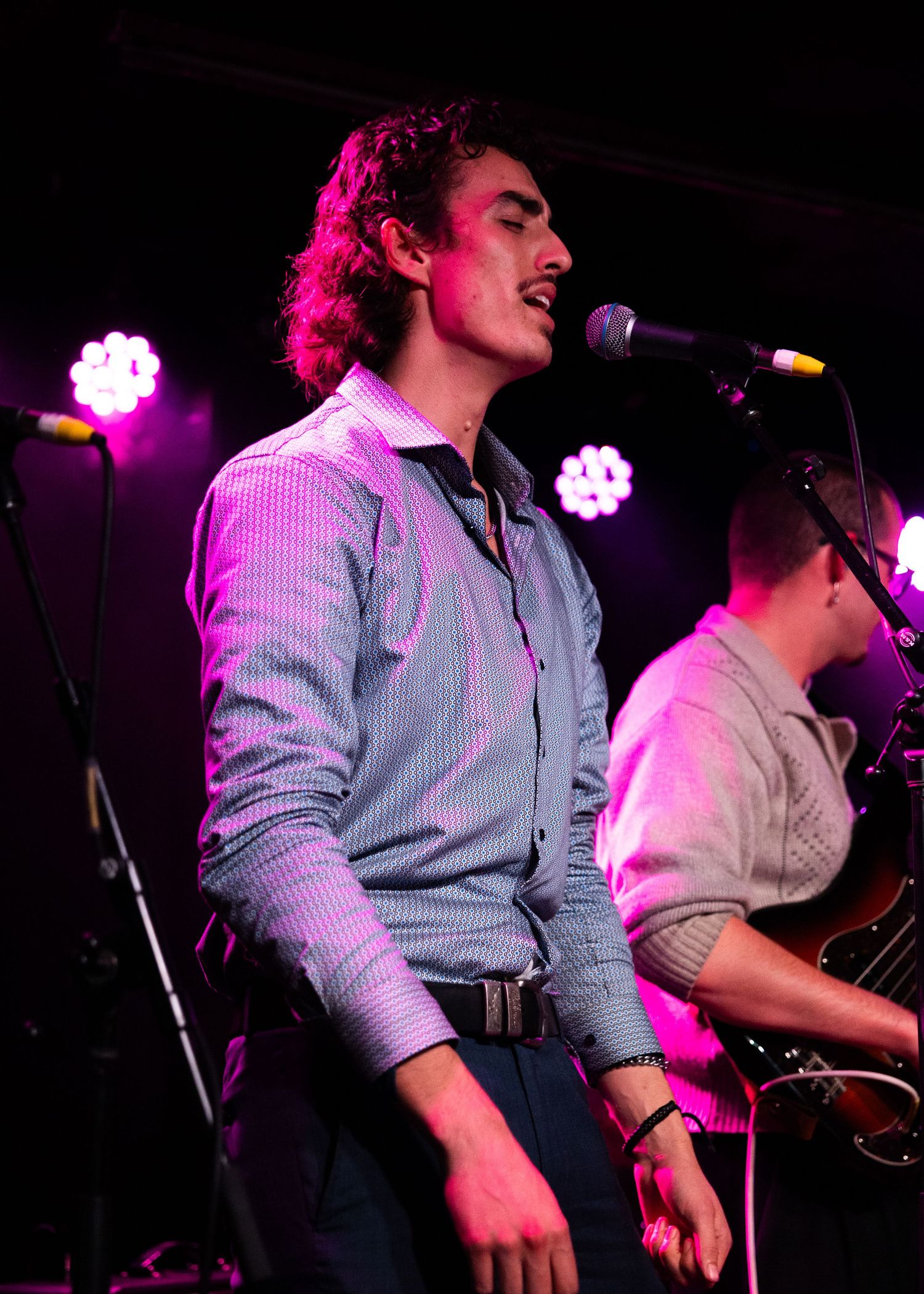 Young male vocalist singing into a microphone on stage under colorful purple stage lights.