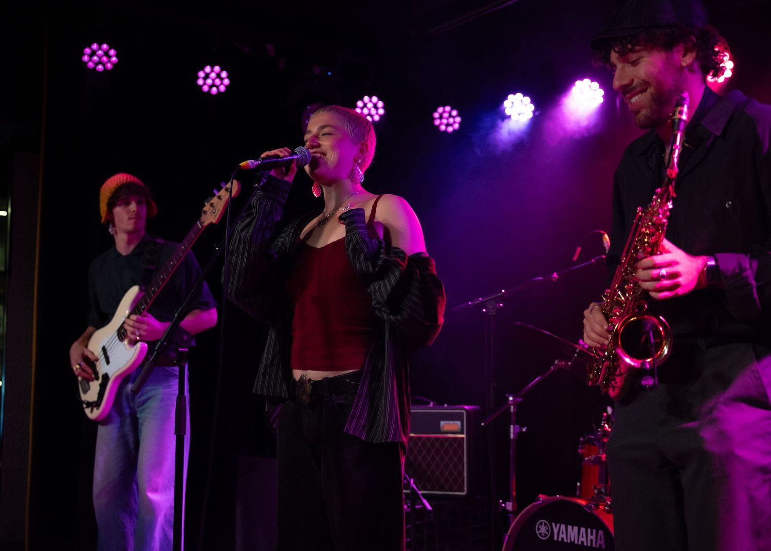 Live band performing on stage with guitarist, female vocalist, and saxophonist under colorful purple and pink stage lights.