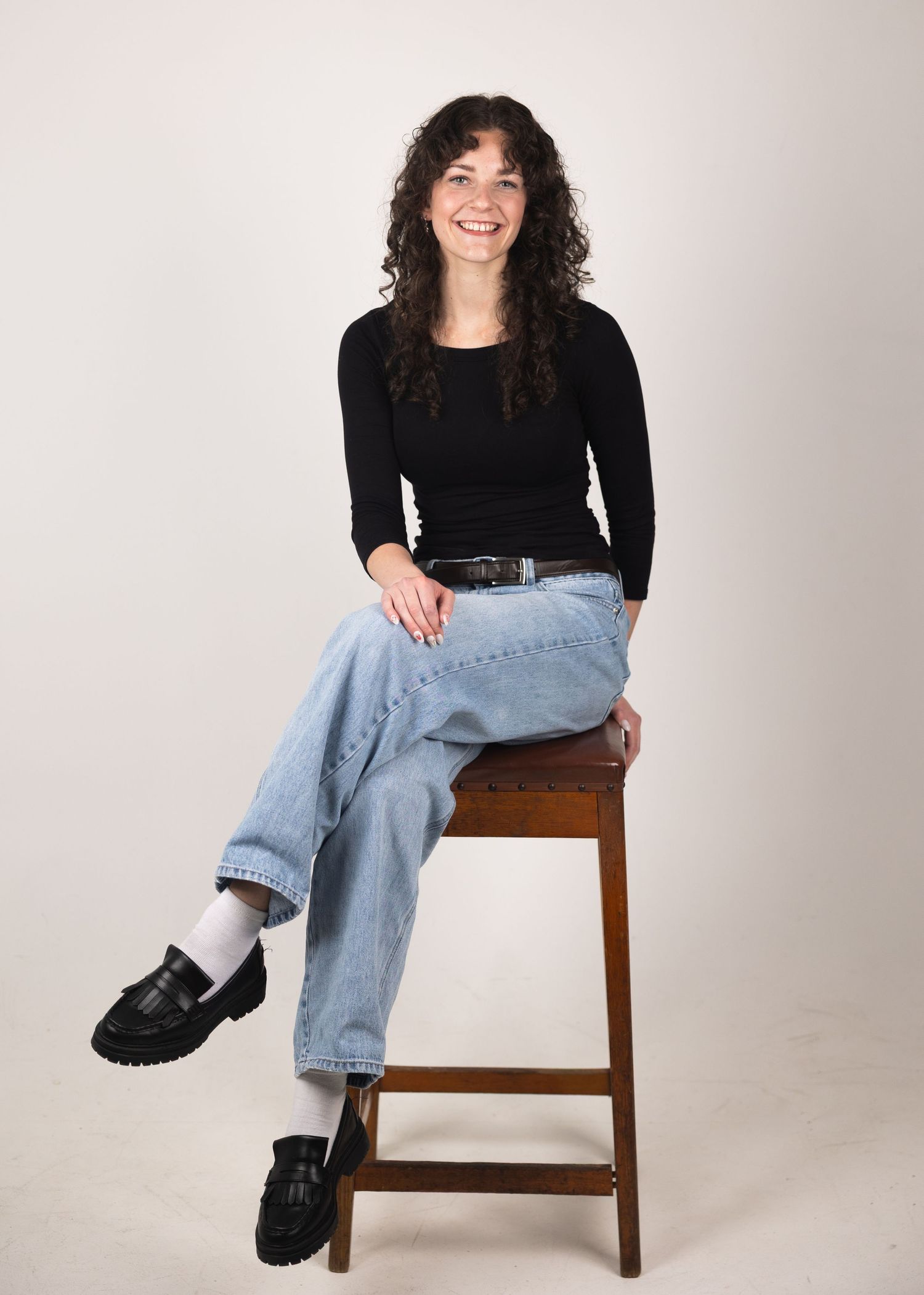 Smiling young woman with curly hair sitting on a wooden stool, wearing black top, light jeans, and black loafers.