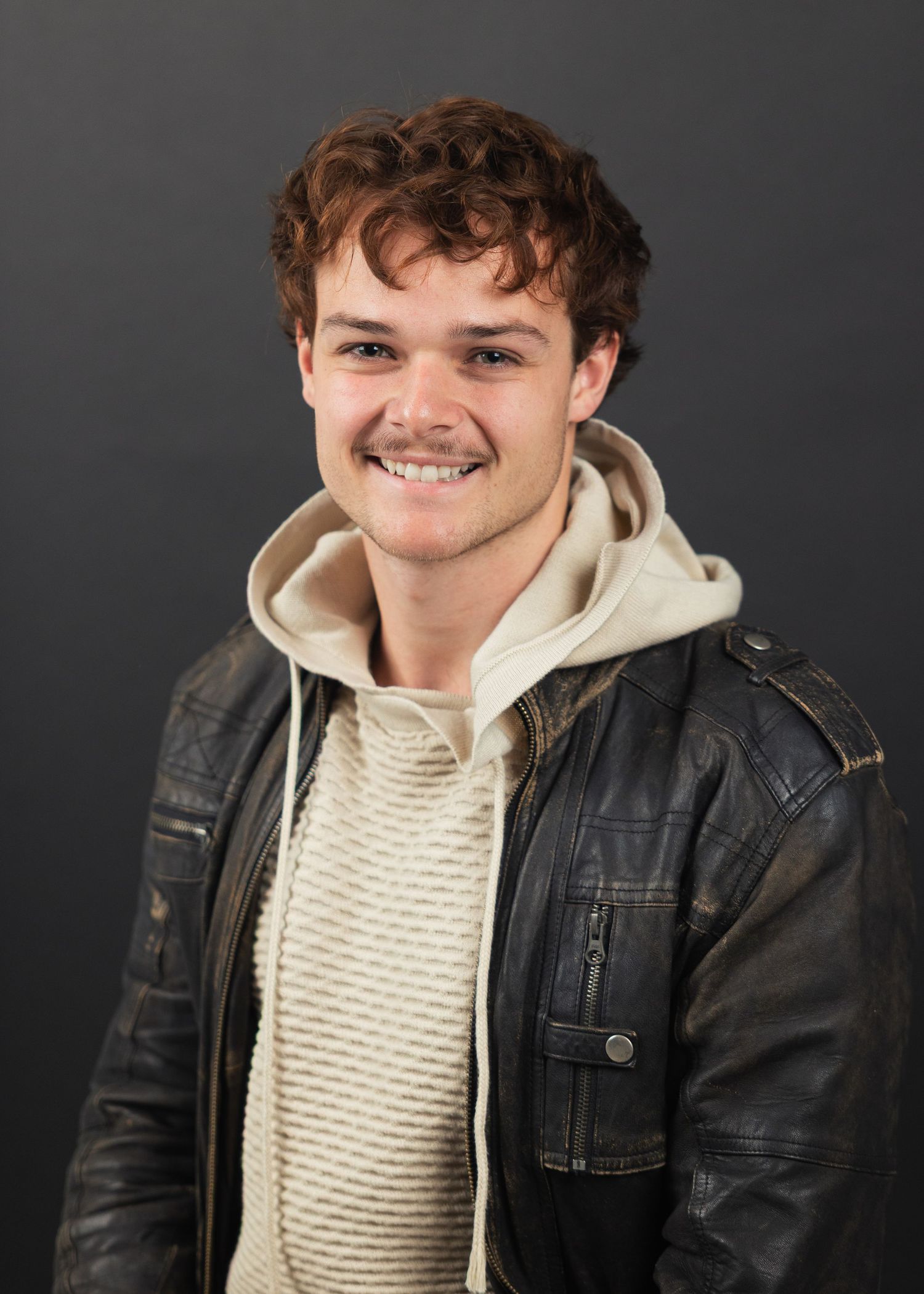 Smiling young man with curly hair wearing a leather jacket and cream hoodie against a gray background.