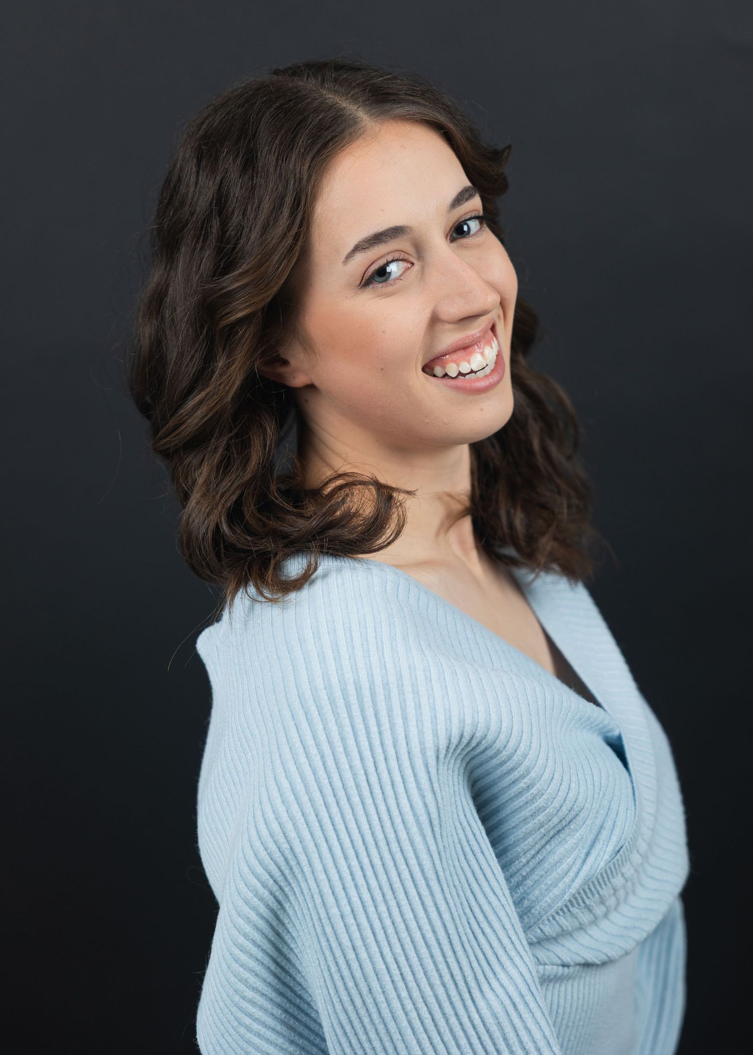 Young woman with dark wavy hair smiling over shoulder in light blue ribbed sweater against dark background.