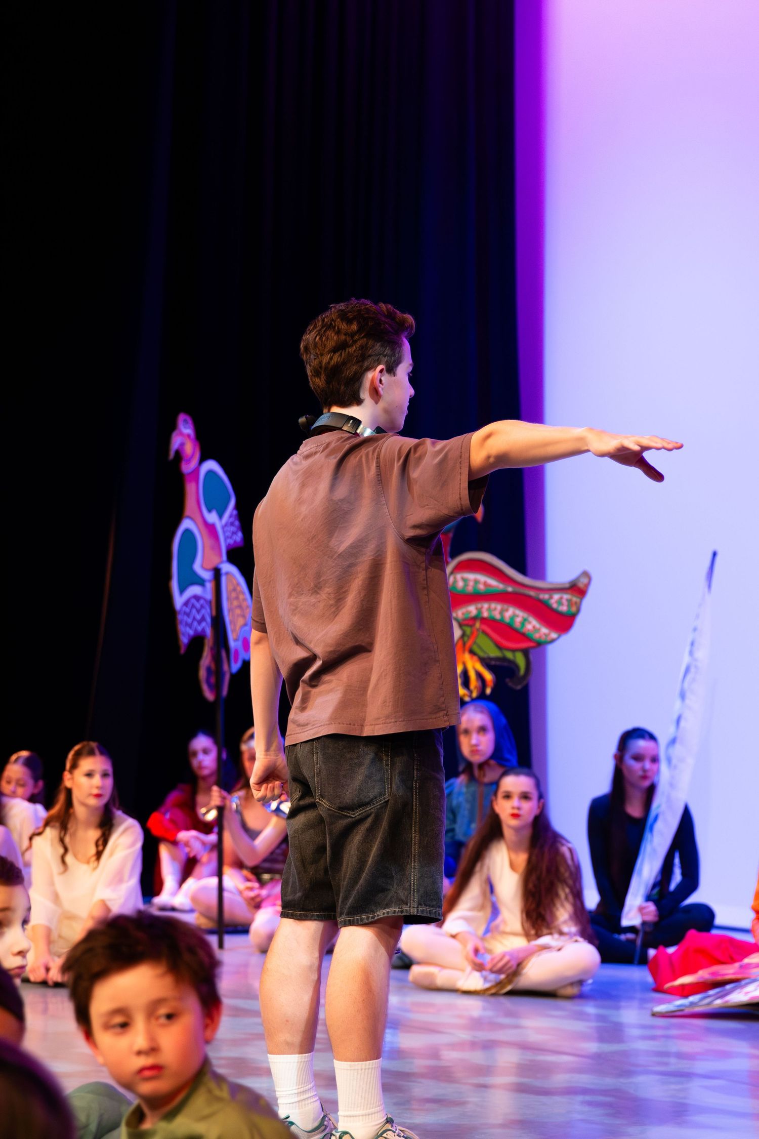 Boy on stage pointing outward during a school theater performance with seated students watching in the background.