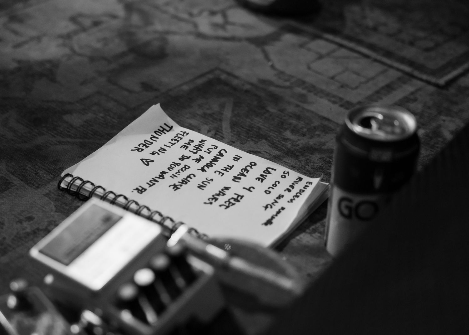 Black and white photo of a handwritten note on a spiral notepad beside a soda can and electronic device on a map.