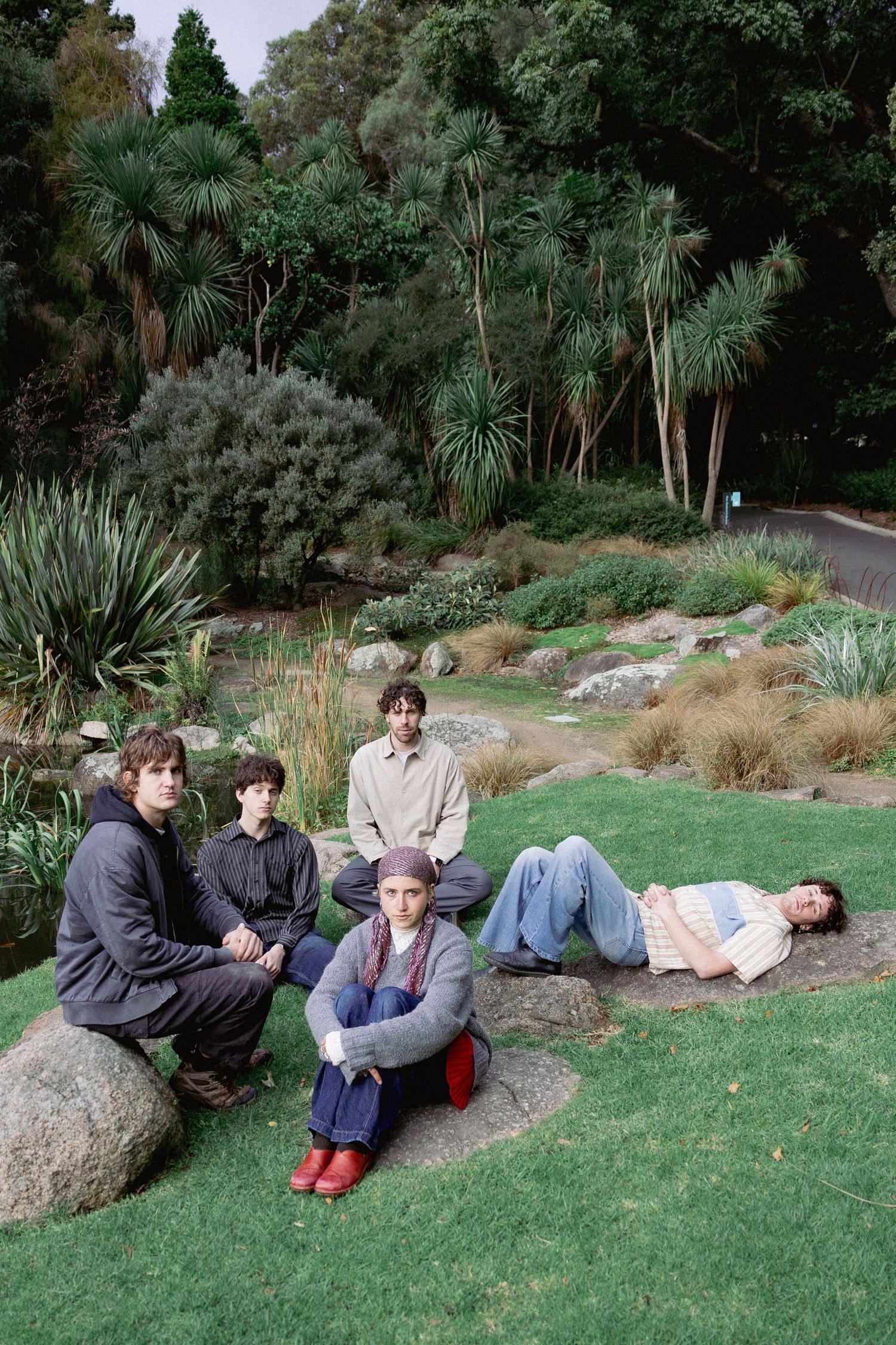 Five young people relax on rocks and grass in a lush botanical garden with diverse plantings.