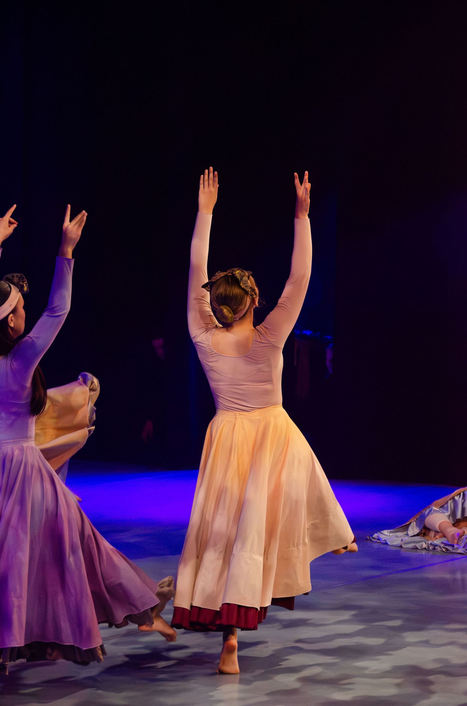 Dancers in flowing white and purple dresses perform graceful movements on a dimly lit stage with blue lighting.