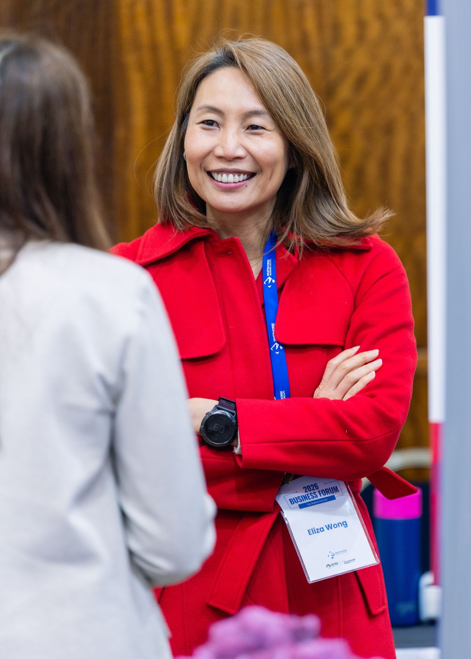 Woman in red jacket with name tag smiles at a professional conference event with arms crossed.