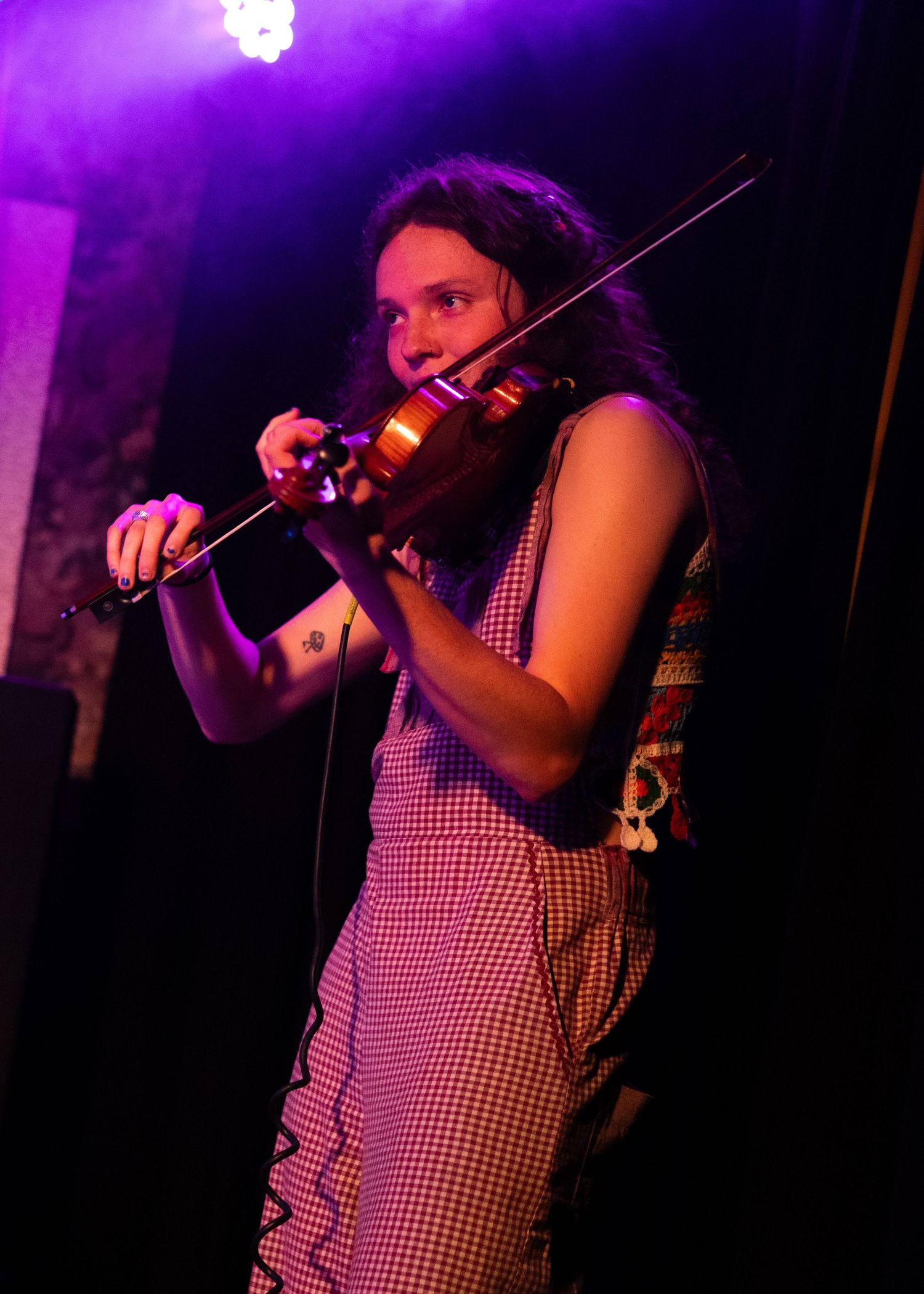 Woman playing violin on stage under purple stage lighting, wearing a checkered dress with a tattoo on her arm.