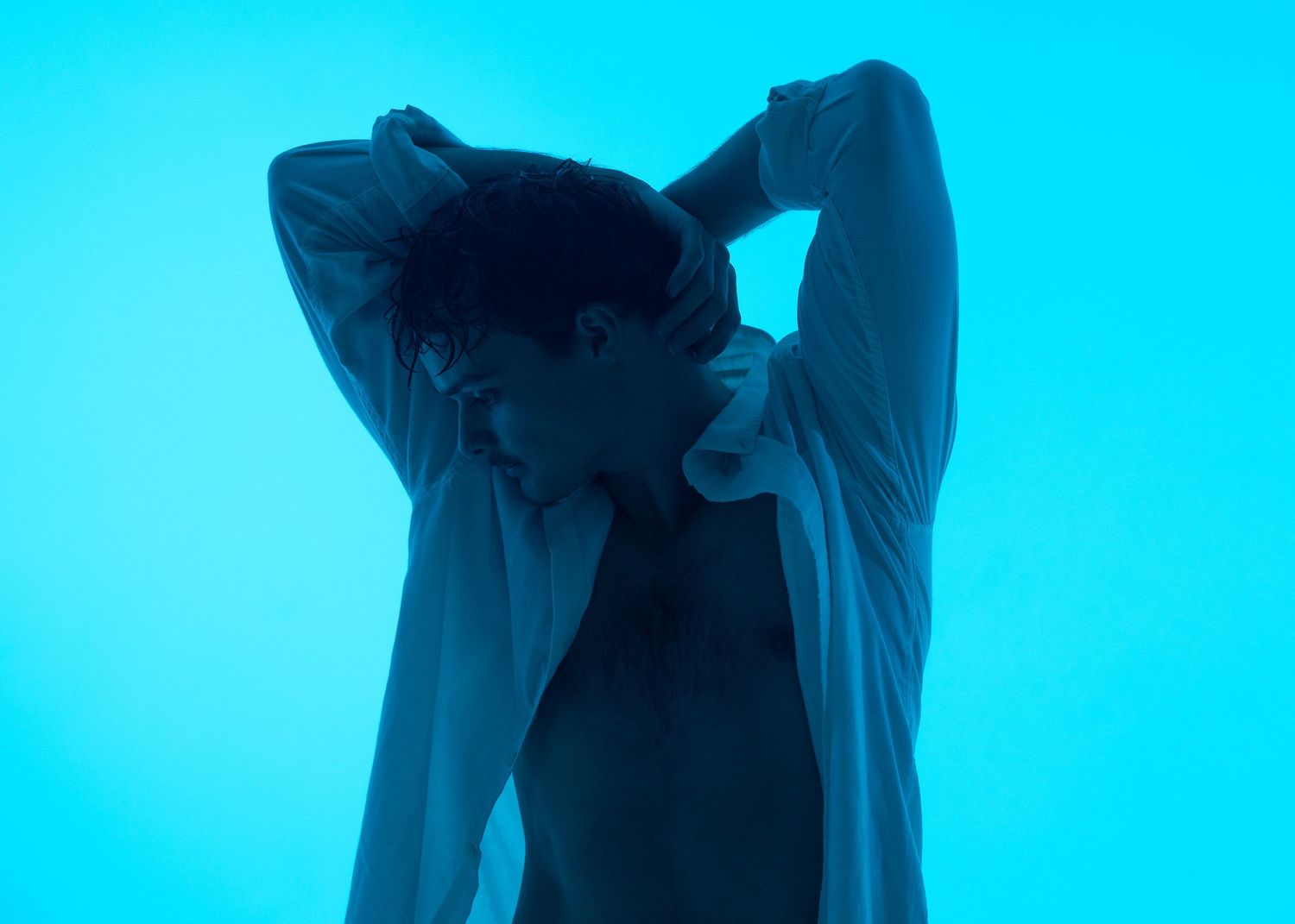 Young man posing with hands behind head, wearing an open white shirt against a vibrant blue background.