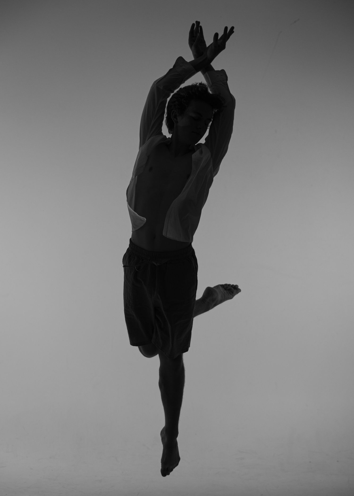 Black and white photo of a male dancer mid-leap, arms crossed overhead, one leg raised, showcasing grace and athleticism.