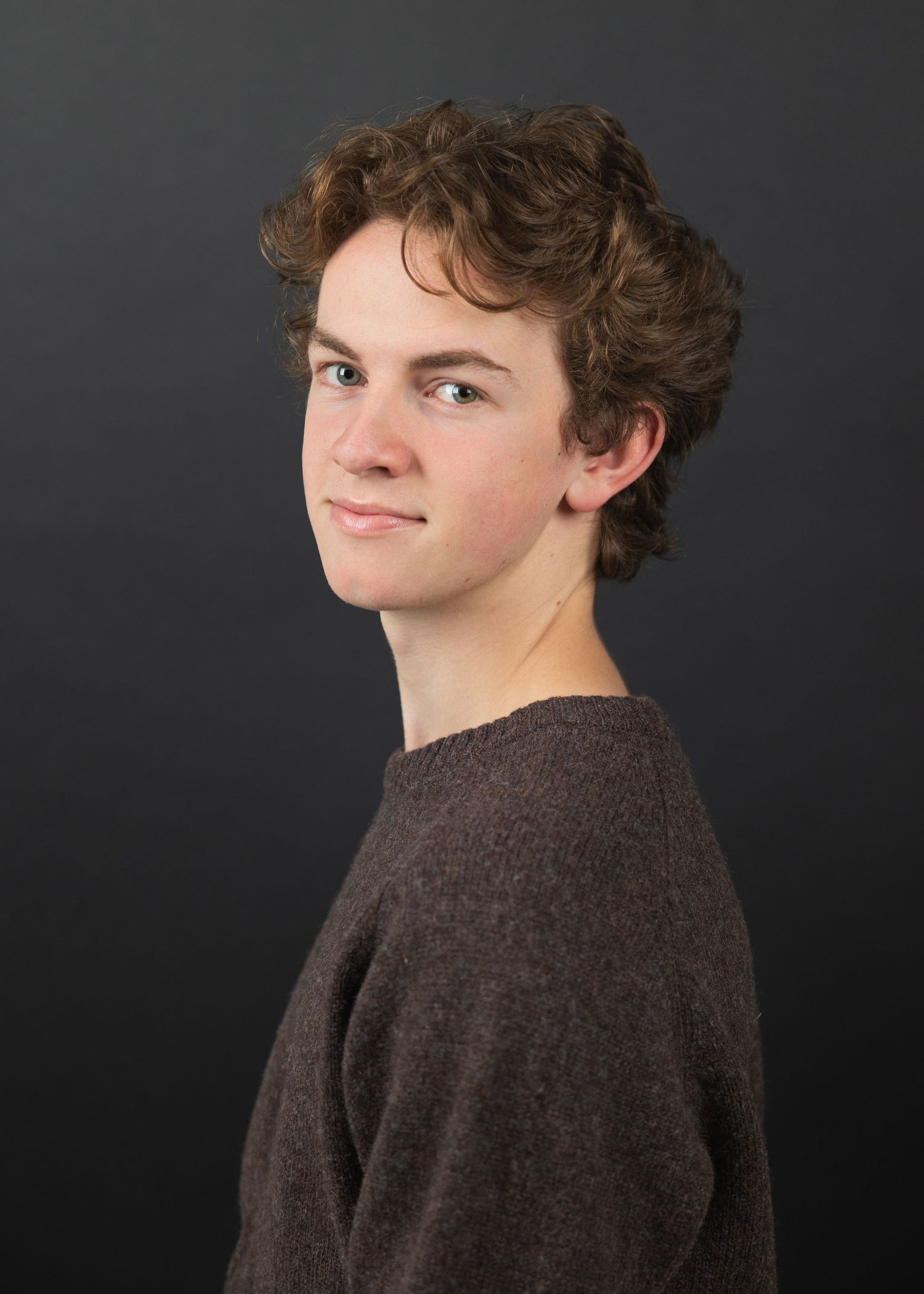 Young person with curly brown hair wearing a brown sweater, posing against a dark gray background.