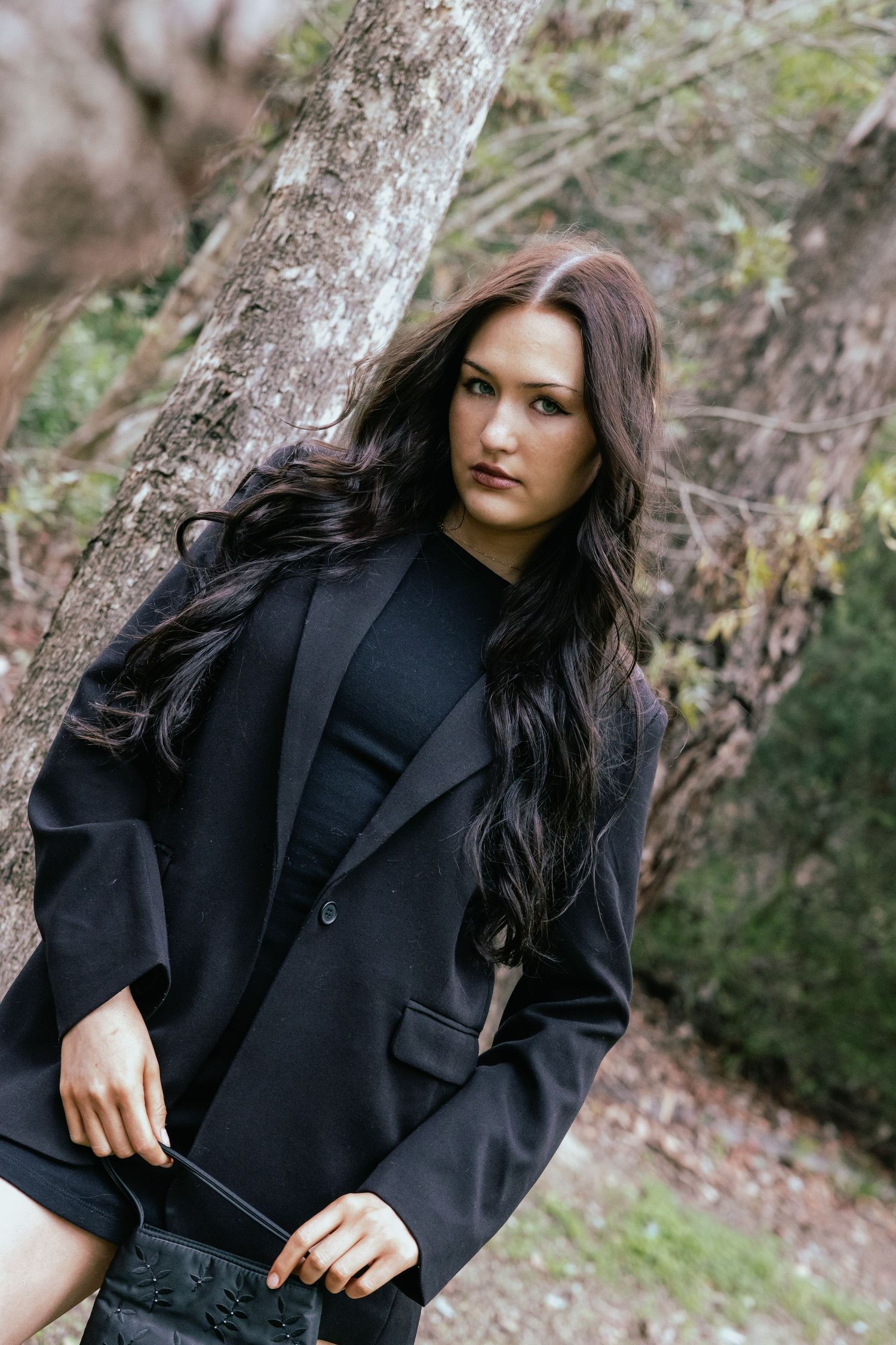 A young woman with long dark hair poses in a black blazer among trees in a moody outdoor forest setting.