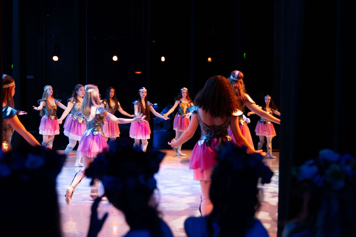 Young girls in colorful tutus perform a dance recital on a stage with dramatic lighting before an audience.