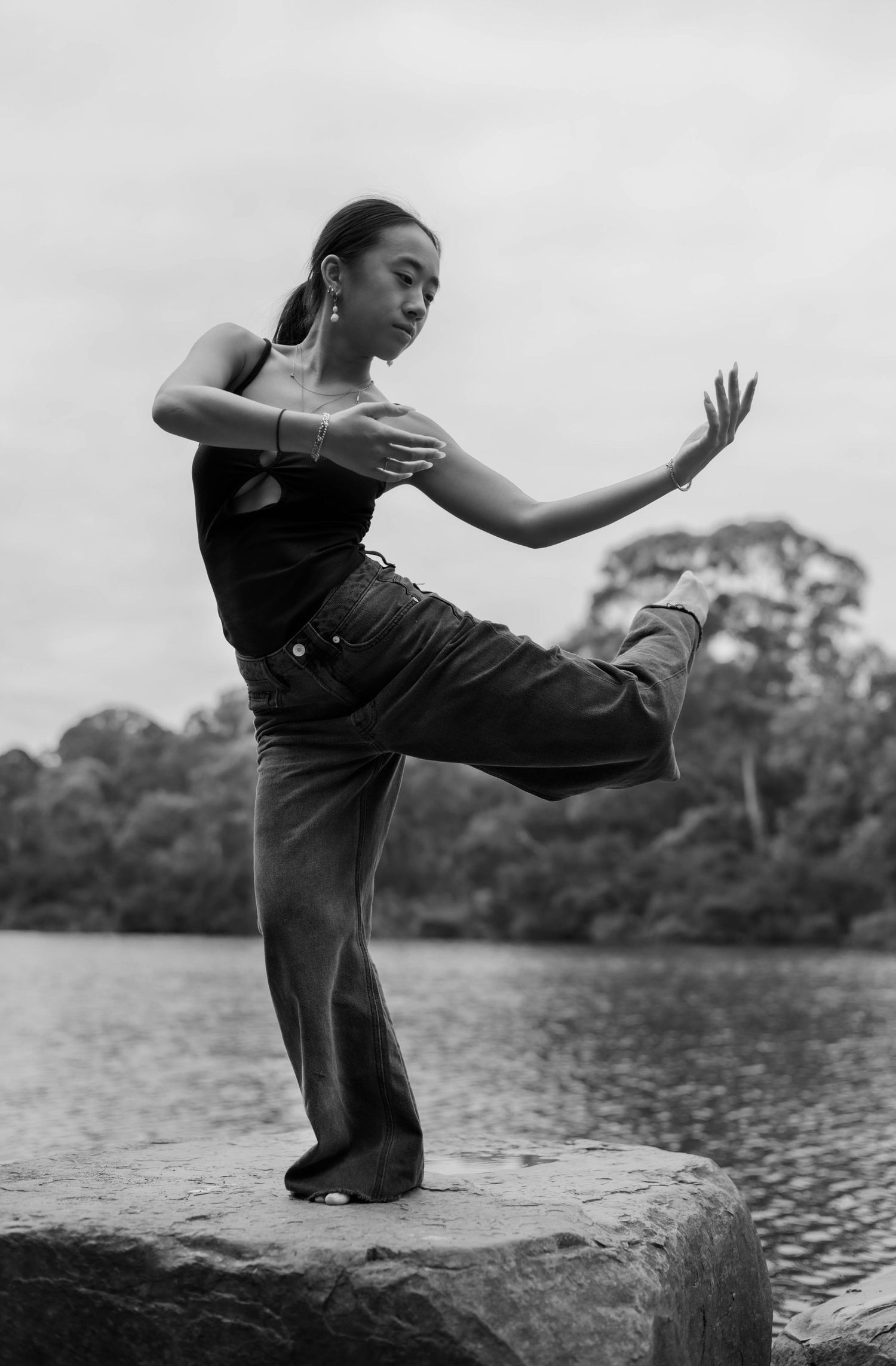 A dancer in overalls performs an elegant pose on a rock by a lake in a black and white artistic photograph.