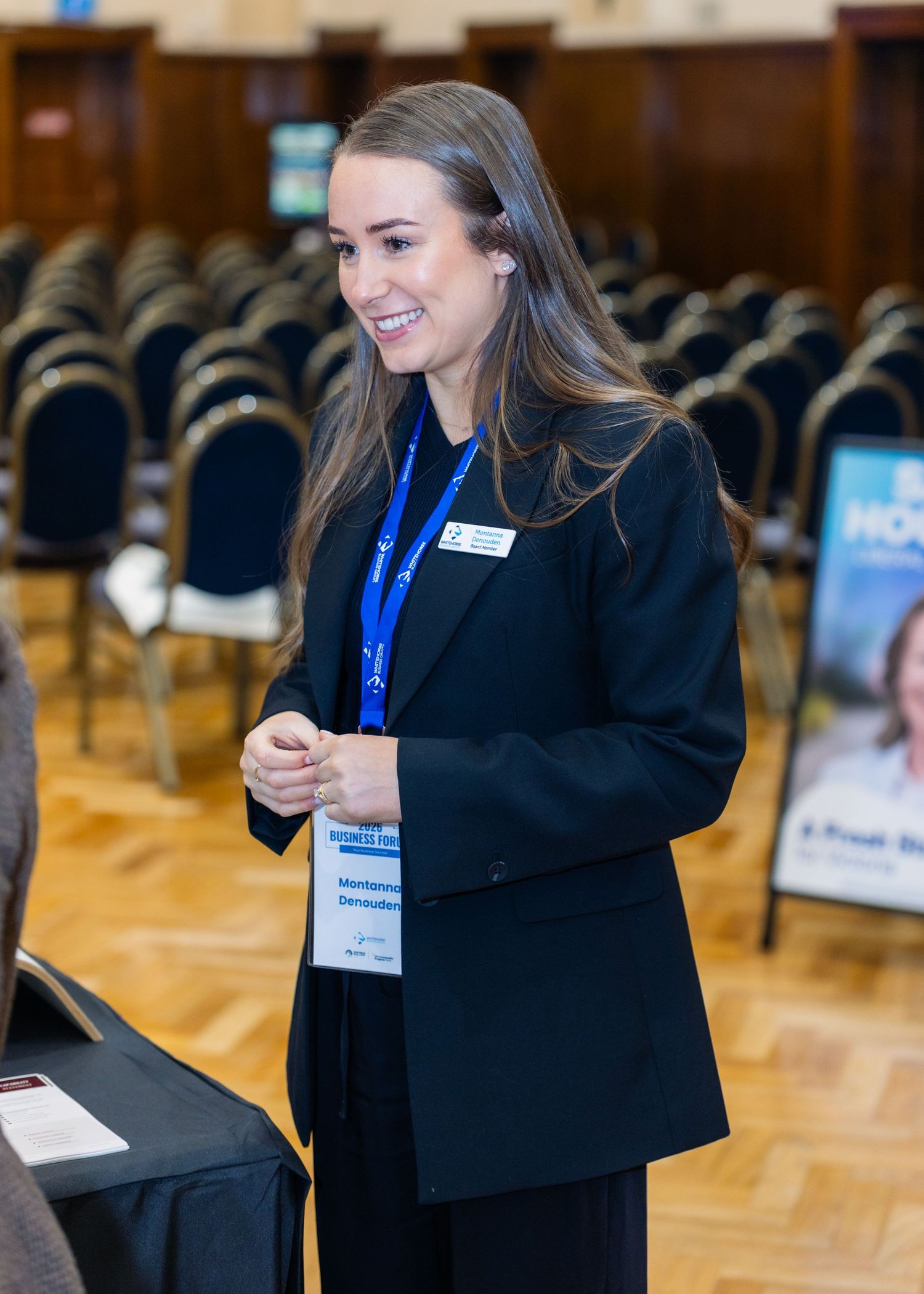 A smiling woman in a black blazer with a blue lanyard stands at a professional conference event.