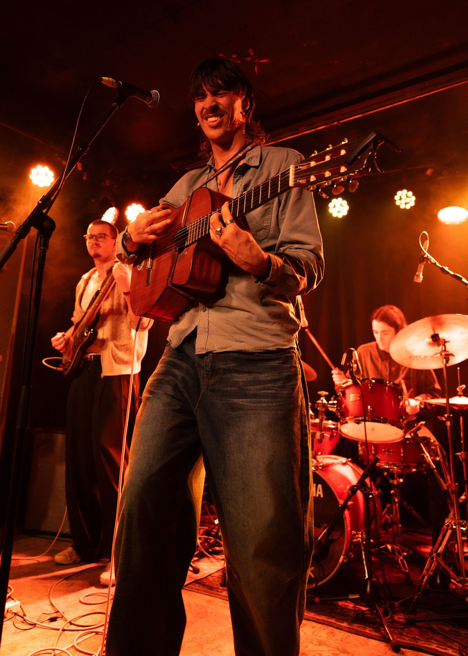 Live band performance with guitarist smiling on stage, drummer and bassist visible in warm amber lighting.