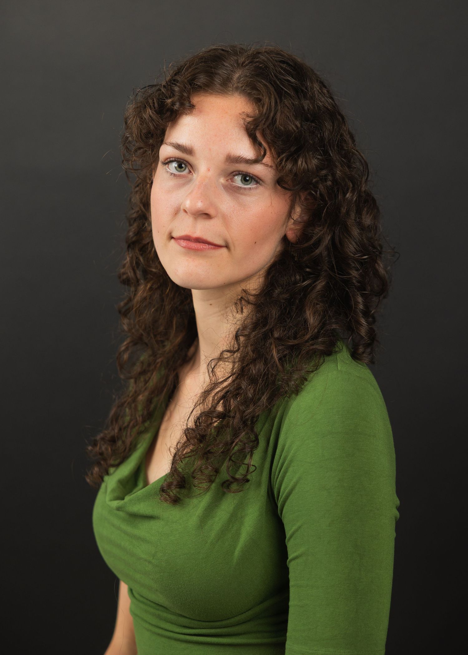 Young woman with curly brown hair wearing a green top poses against a dark background.