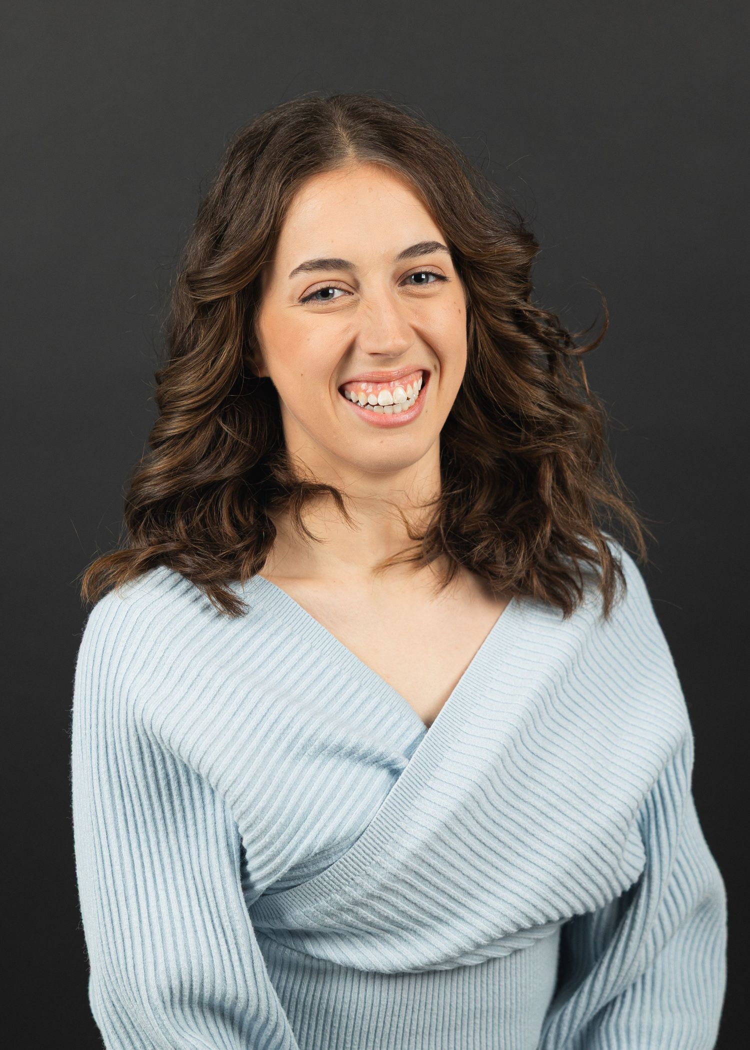 Smiling young woman with curly brown hair wearing a light blue ribbed wrap sweater against a dark background.