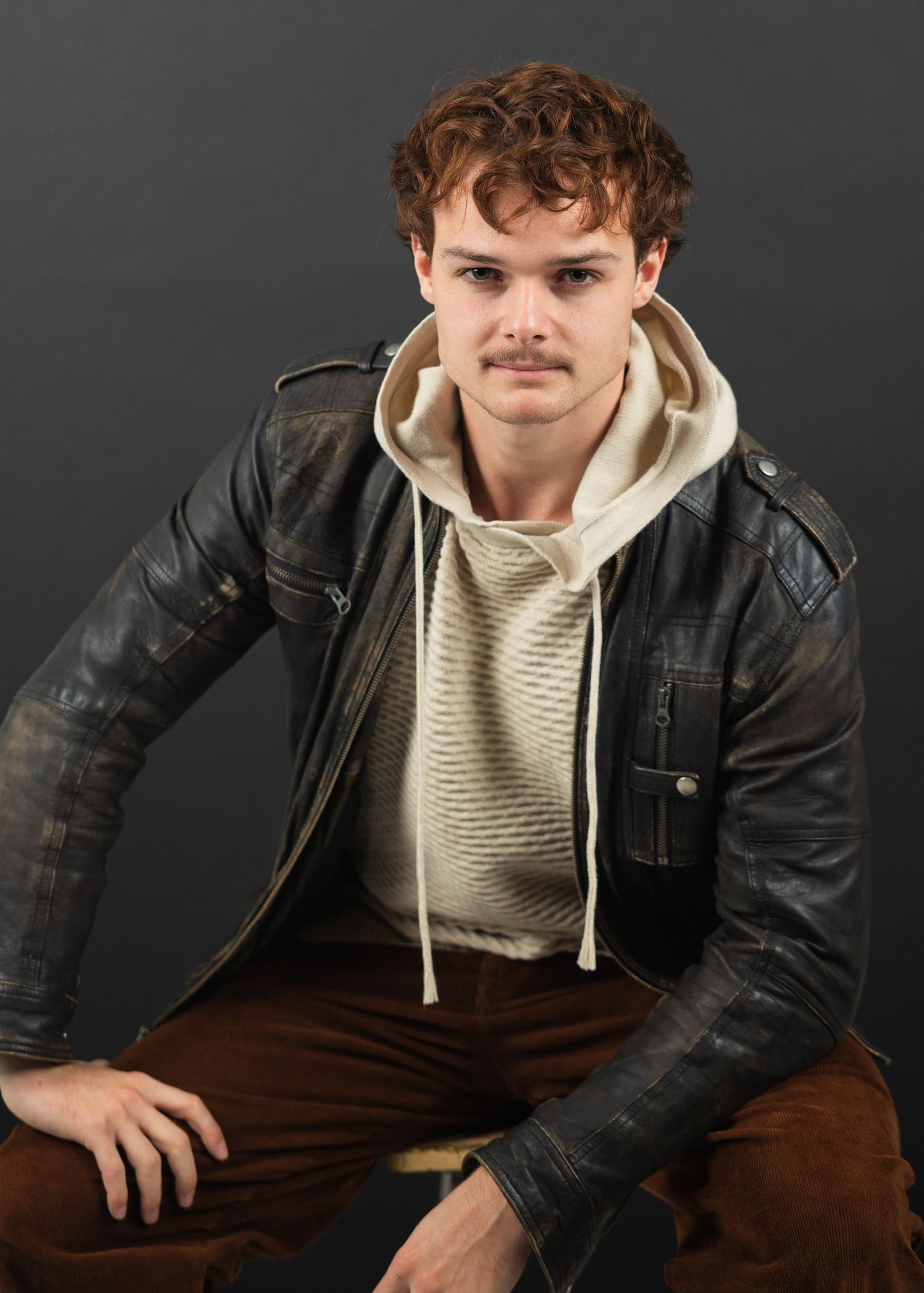 Young man in distressed leather jacket and cream hoodie poses against dark gray background.
