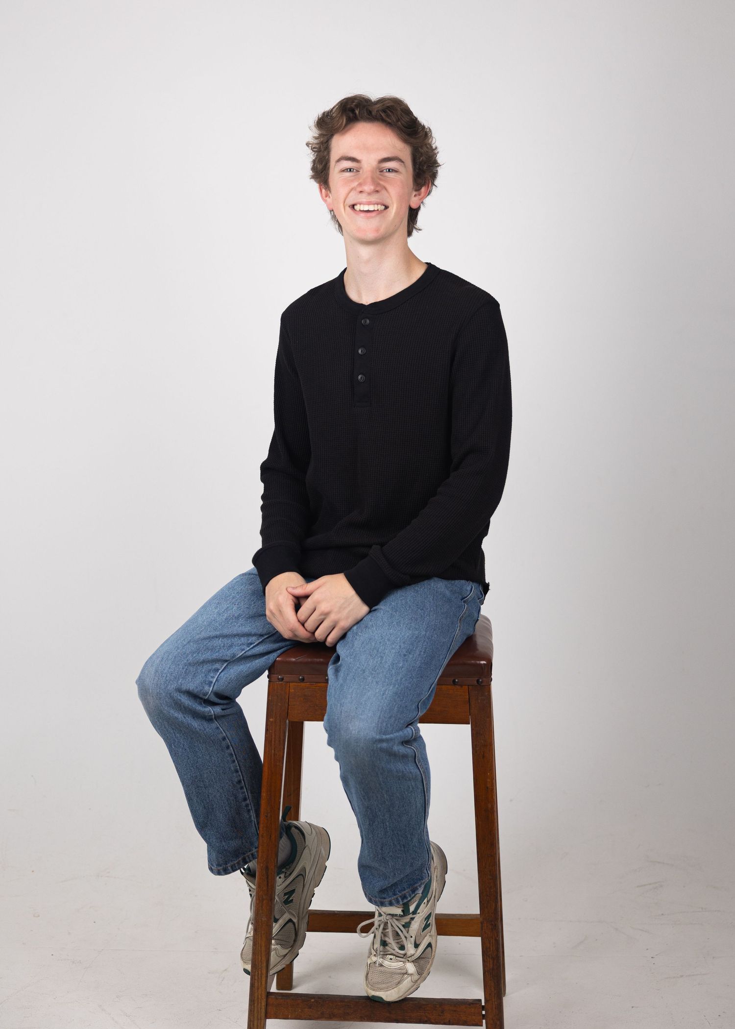 Smiling young man in black henley shirt and jeans sitting on wooden stool against white background.